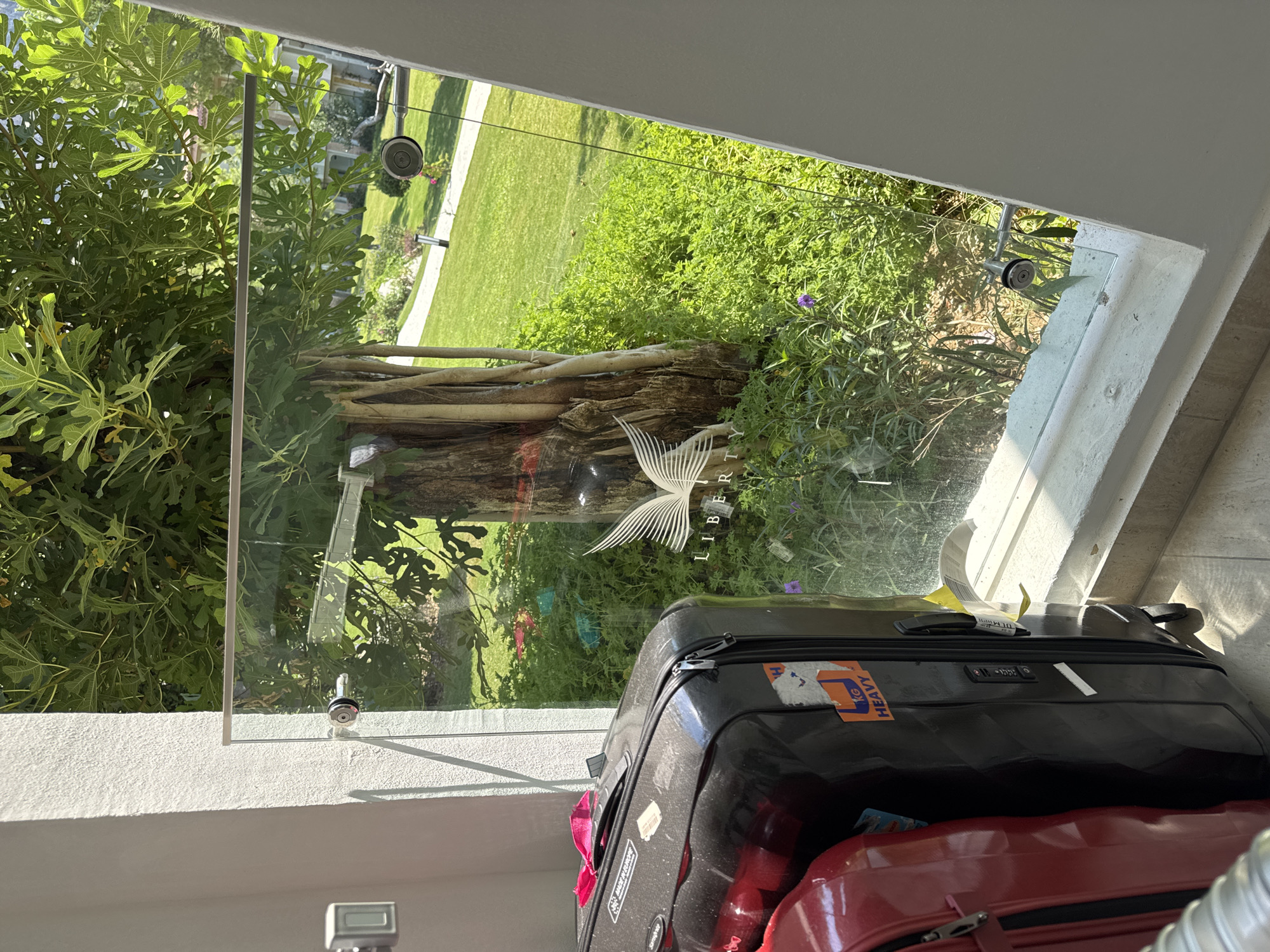 The image shows a view from a balcony in Fethiye, Turkey, looking out through a glass railing. Two suitcases, one black and one red, are on the balcony floor. The black suitcase has several stickers on it, including one that says "MADE IN EUROPE" and another that says "HEAVY". In the background, there is a lush green lawn, a winding path, and trees with large leaves. The sun is shining, casting shadows. There is a logo of a bird with outstretched wings on the glass railing.