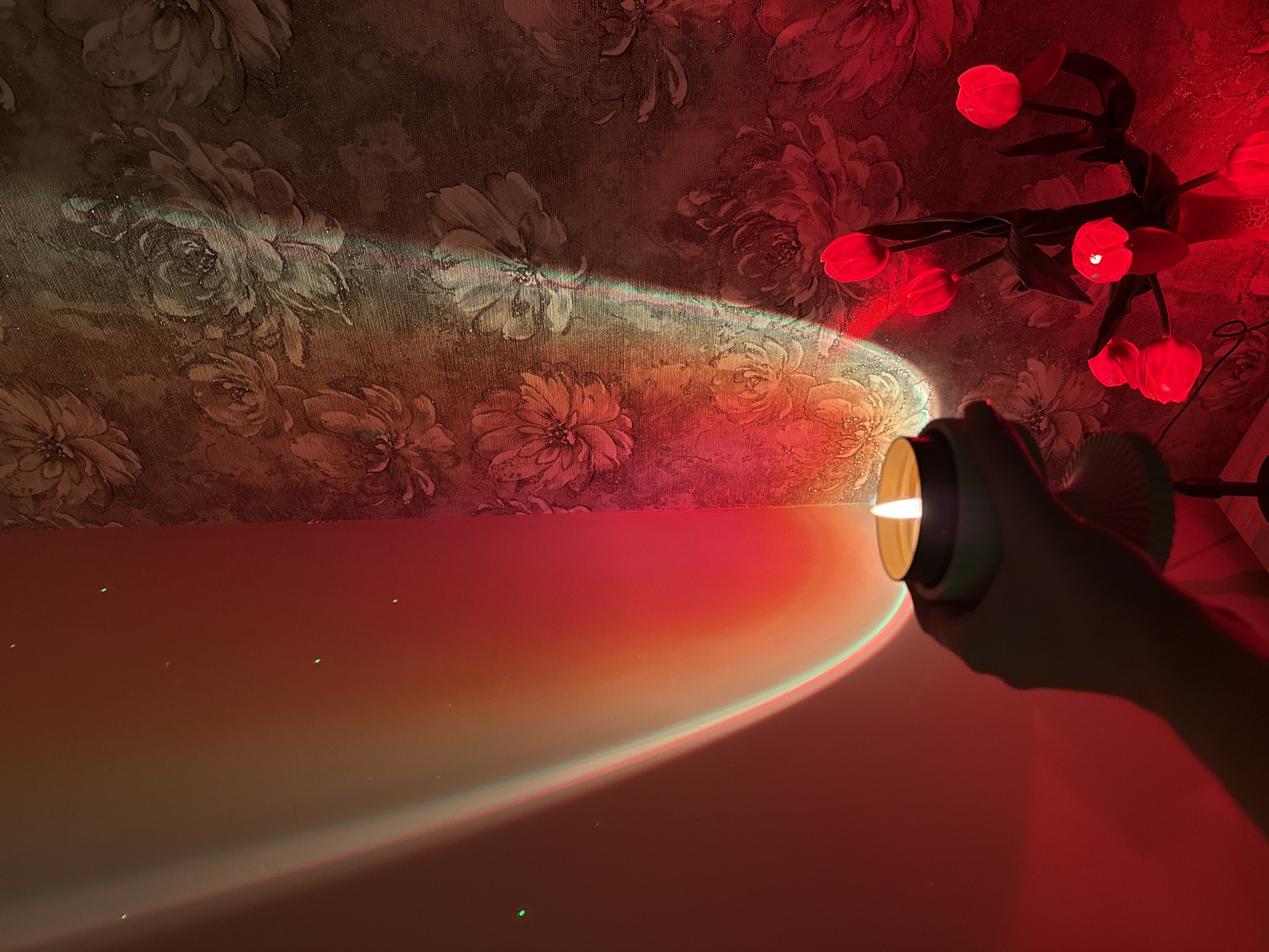 An unseen person's hand holds a lit candle in a light-colored holder. The candle is positioned in the lower center of the frame. A gradient of light, transitioning from red to orange and yellow, is cast on the left wall. On the right wall, a floral wallpaper is illuminated by red light and a narrow rainbow-colored beam. In the lower right corner, a red light fixture shaped like tulips is visible. The overall lighting creates a warm and atmospheric scene.
