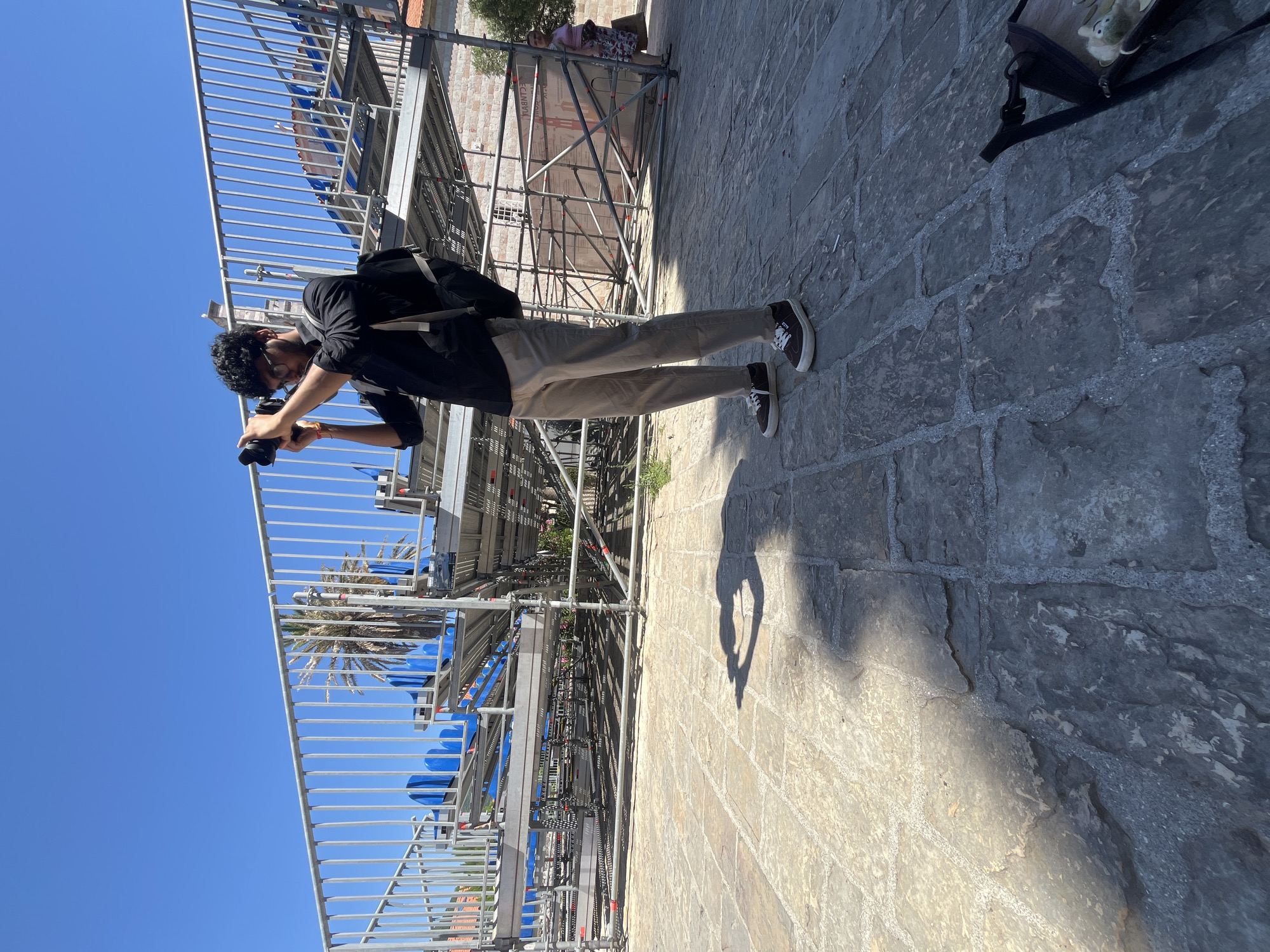 In Budva, Montenegro, a young man, dressed in a black button-down shirt, light-colored trousers, and dark sneakers, stands on a paved stone surface, actively photographing a temporary structure made of scaffolding and seating. He is wearing glasses and has a backpack slung over his shoulders. Behind him, the scaffolding supports rows of blue seats, suggesting an outdoor event venue. The sky is clear blue, and a palm tree is visible in the background. The sunlight casts a shadow of the man onto the stone ground. Further in the background, other structures and people are visible, indicating an urban environment.