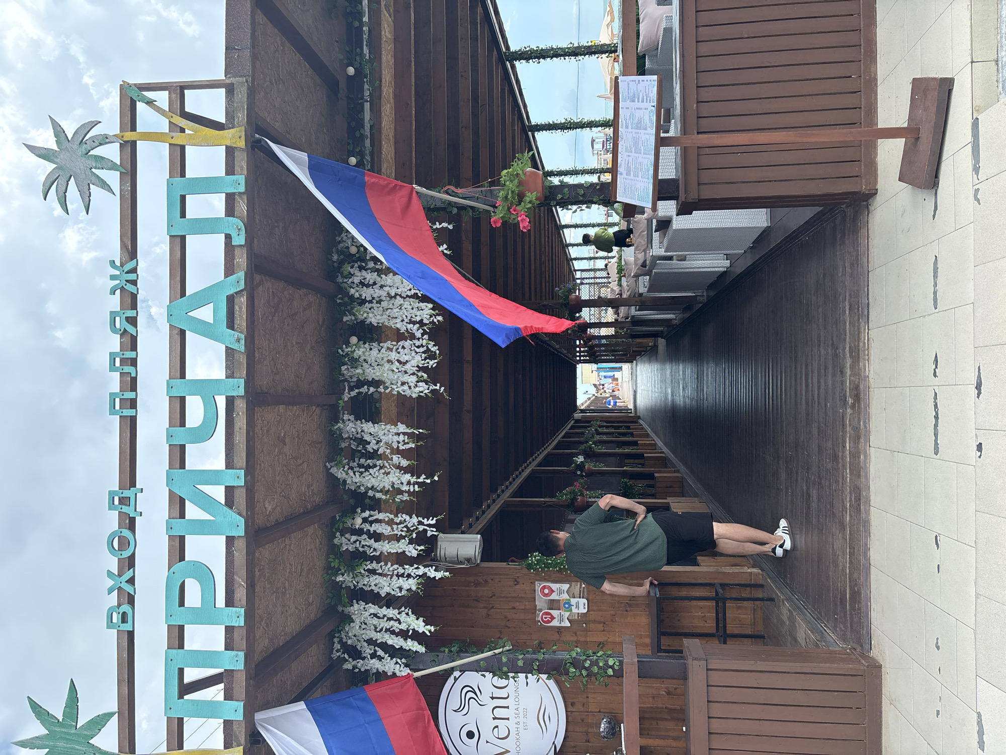 The image captures the entrance to a beach establishment in Sochi, Russia, identified by a sign reading "ВХОД В ПЛЯЖ ПРИЧАЛ" which translates to "ENTRANCE TO THE BEACH PIER". A man in a green striped shirt and black shorts stands with his hands on his hips, facing away from the camera. To his left, a Russian flag is displayed. The establishment features a wooden walkway leading into an open-air seating area, with tables and chairs visible. White floral garlands hang from the wooden ceiling. In the background, people are seen walking towards the beach, and the sea is visible. The weather appears overcast.