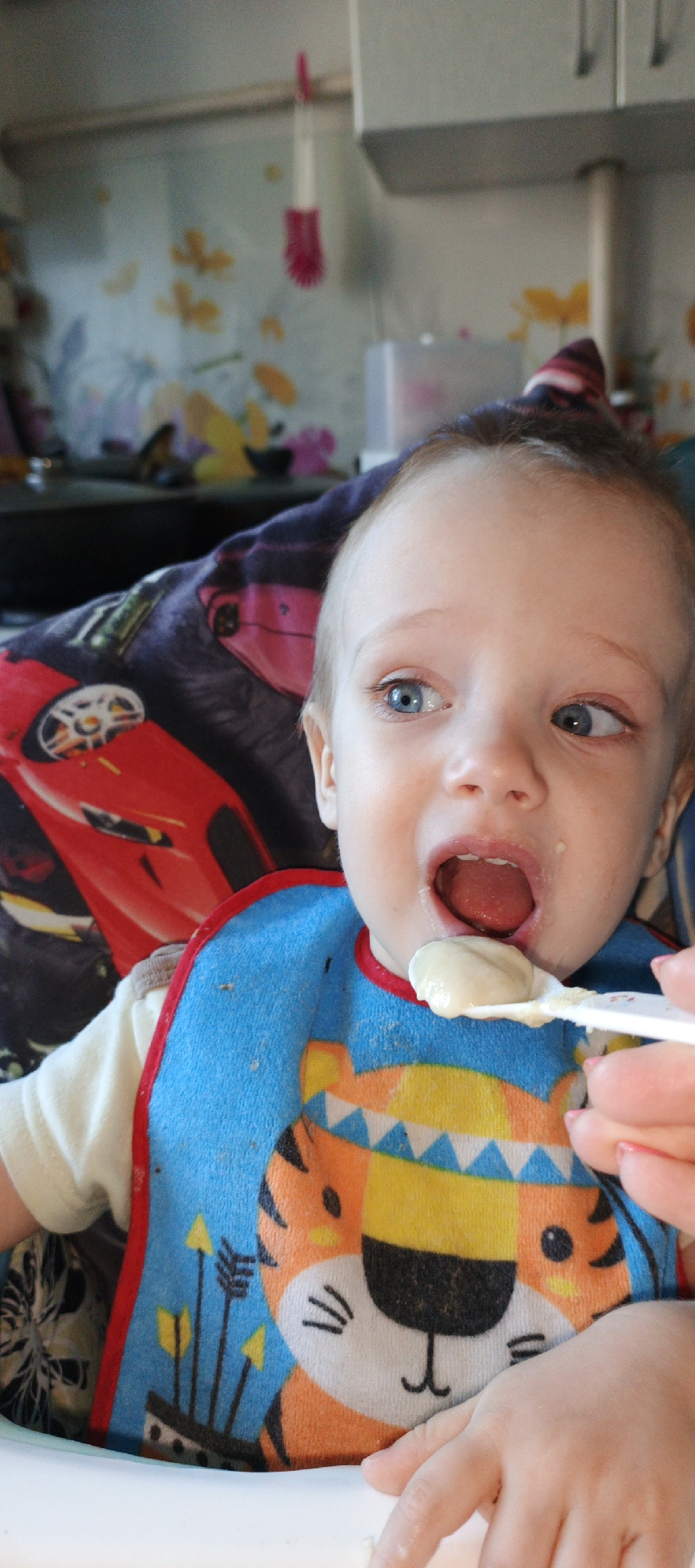 The image depicts a baby with blue eyes being spoon-fed. The baby is wearing a blue and orange bib with a tiger design and is seated in a highchair with a car-themed cover. A hand is holding a spoon with a beige food substance, poised to feed the baby. The background shows a kitchen with a floral patterned wall and white cabinets. Based on other available knowledge, this image likely came from the city of Chaikovsky, Russia.