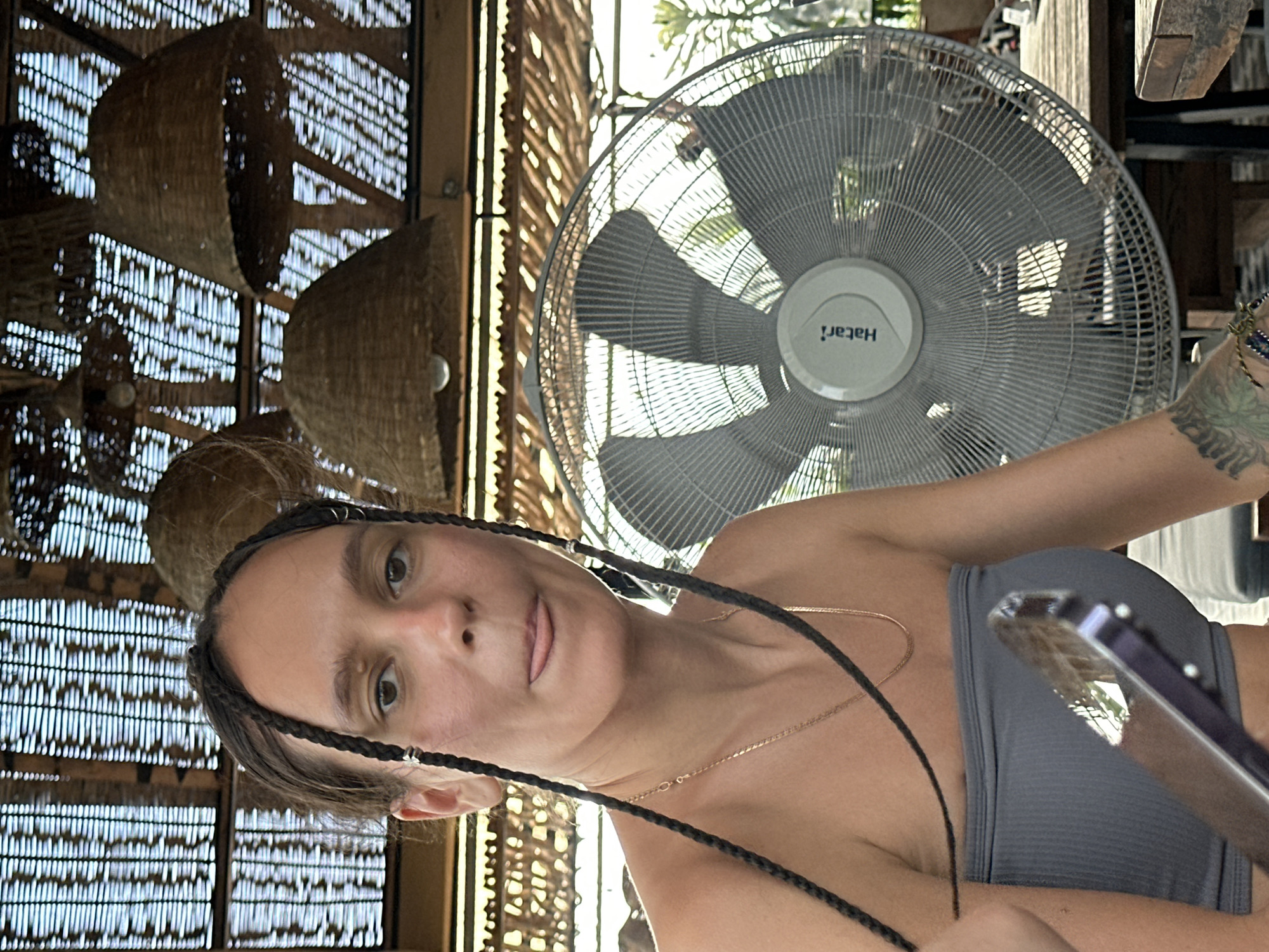 A woman is featured in a close-up, rotated portrait, appearing to take a selfie or be photographed in what looks like a cafe, restaurant, or covered outdoor area in Hà Giang 1 Ward, Vietnam. She has dark hair styled in several braids, a light complexion, and is wearing a dark grey strapless top and a thin gold chain necklace. Her expression is playful, with her tongue slightly extended.
To her right, a white Hatari brand standing or ceiling fan is prominent, its blades visible behind a protective grille. Multiple woven, basket-like light fixtures or decorative elements hang from the ceiling in the background. The setting features light filtering through a patterned, possibly bamboo or rattan, ceiling or wall structure, suggesting a rustic or tropical aesthetic. Bright natural light illuminates the scene, indicating it's daytime. A tattooed arm is visible on the right, holding a reflective object, likely a smartphone. The overall impression is casual and relaxed.