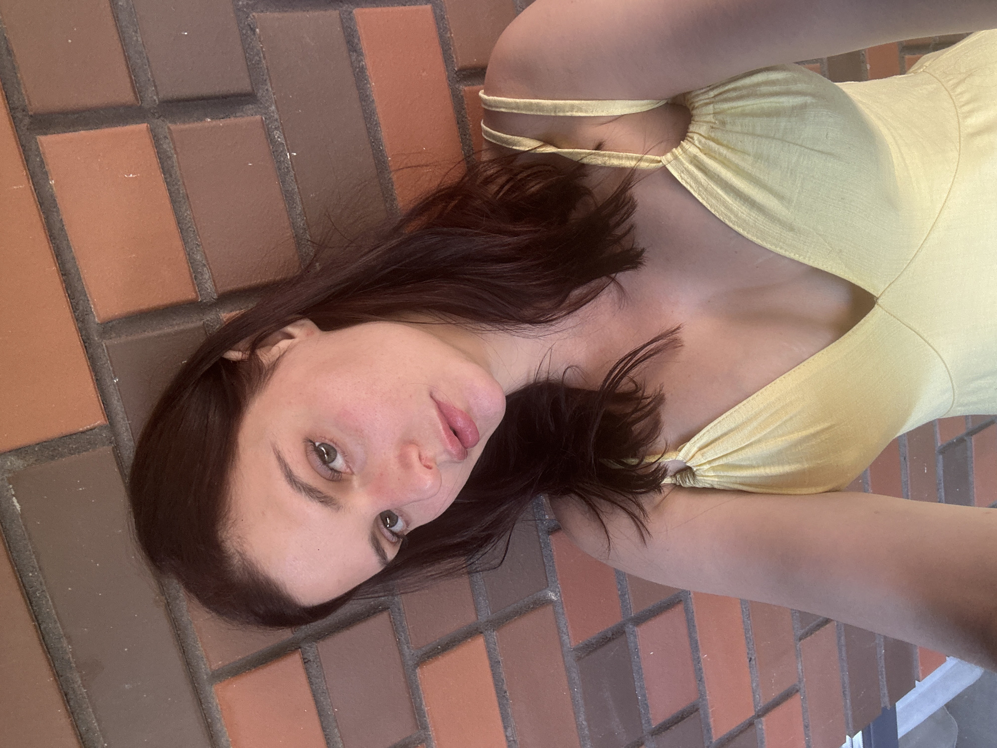 A young woman with dark hair and fair skin is leaning against a brick wall, looking into the camera. She is wearing a yellow dress with thin straps. The brick wall is made up of reddish-brown bricks with gray mortar. The lighting suggests an outdoor setting during the daytime.