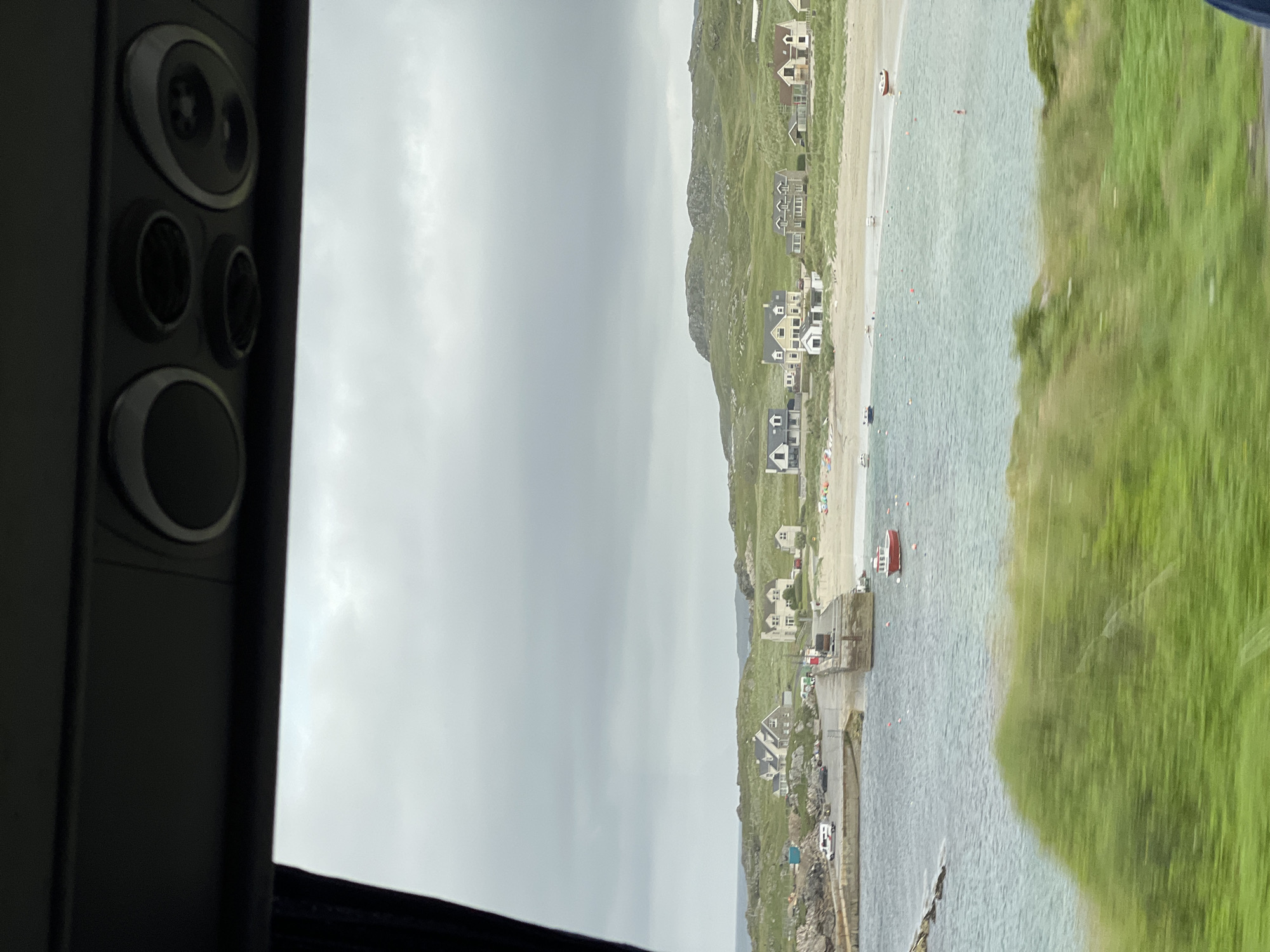 This media file depicts a picturesque coastal scene at Lagnalore, Ireland. The image is oriented vertically, with the left side dominated by a dark, textured surface that appears to be part of a vehicle's interior, possibly a window frame or dashboard, featuring several circular elements.
The main subject is a scenic view of a bay with a sandy beach, turquoise water, and a backdrop of grassy hills dotted with houses. The houses are varied in style, with some appearing traditional and others more modern. A small pier or jetty extends into the water, with a red boat moored alongside it. Several other small boats and buoys are visible scattered across the water.
The beach has a few scattered people, suggesting a leisurely day, and there are a few parked cars visible along the road near the waterfront. The sky is overcast, indicating a cloudy or possibly humid day. The overall impression is one of a serene, rural coastal settlement, likely in the summertime given the presence of people enjoying the beach and water. No visible text is present in the image.