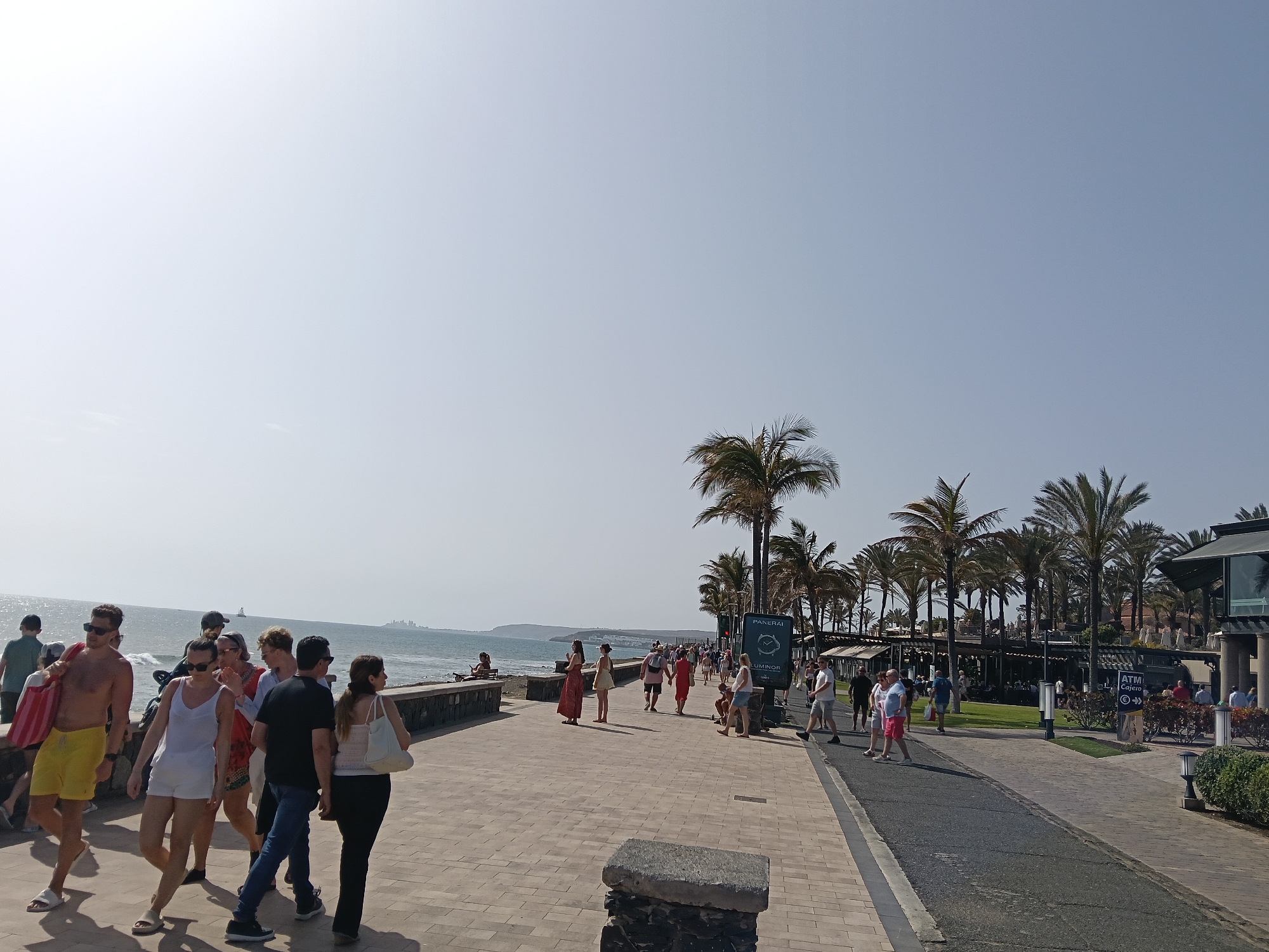 The image depicts a sunlit scene on a paved walkway in San Bartolomé de Tirajana, Spain. A group of people stroll along the walkway, some in beach attire, with the ocean and coastline visible in the background. Palm trees line the walkway, providing shade. A building and signage are visible on the right side of the image.