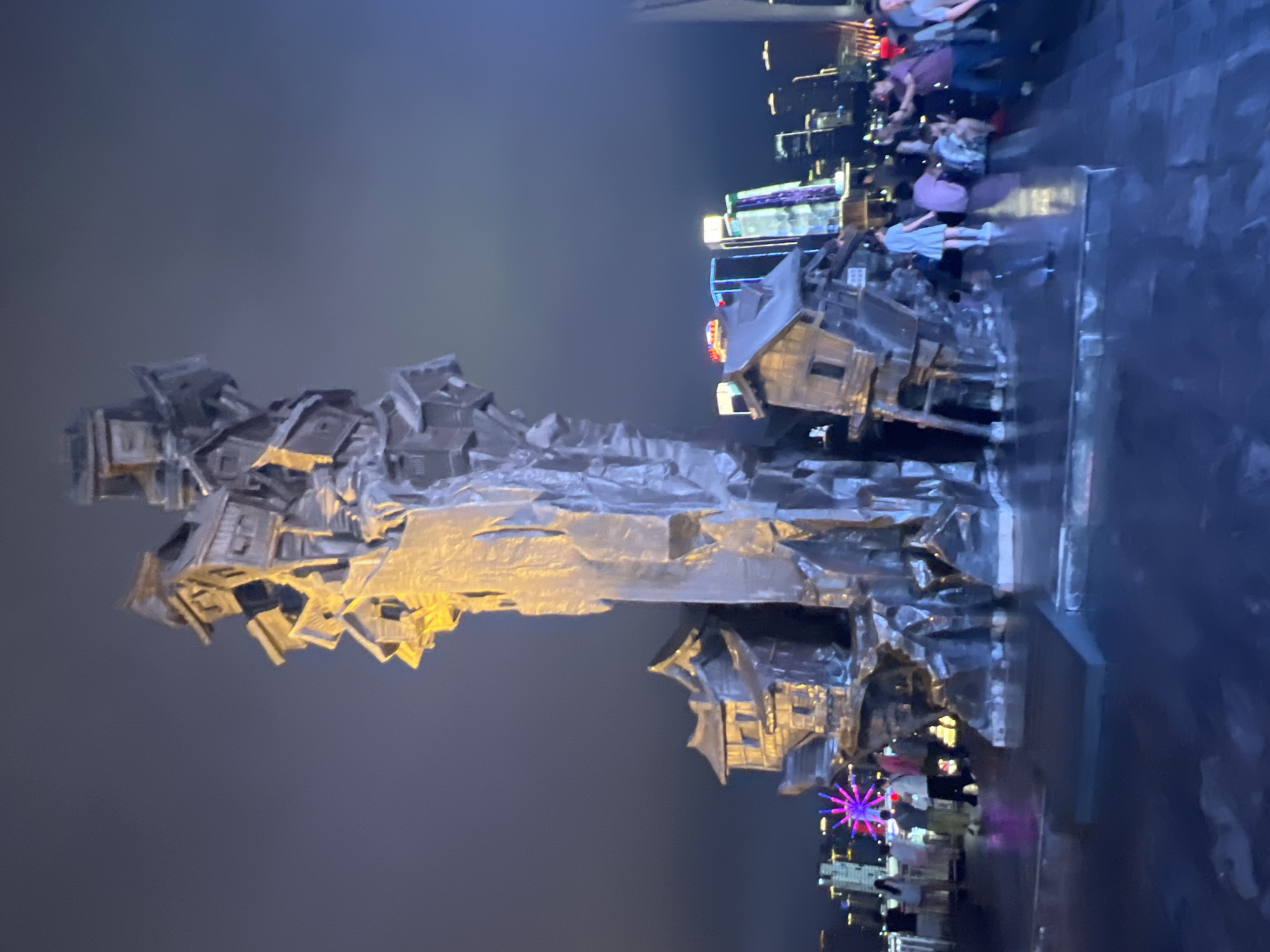 The media file depicts a nighttime scene in Yuzhong District, China. The primary subject is a large, abstract sculpture made of metallic material, appearing to be composed of stacked, fragmented building-like structures. The sculpture is illuminated, with some sections glowing yellow and others reflecting the surrounding lights.
In the background, a cityscape is visible with numerous illuminated buildings and lights, suggesting an urban environment. A Ferris wheel with purple and pink lights is also prominent in the background, further indicating an entertainment or commercial area. Several people are visible in the mid-ground and background, appearing to be gathered or walking, some near the sculpture and others further away. Their presence suggests that this is a public space.
The ground surface, particularly on the right side of the image, appears wet and reflective, possibly indicating recent rain or a water feature. The overall atmosphere is dark, consistent with nighttime, and the lighting suggests a vibrant urban setting. There is no visible text in the image. The scene evokes a sense of urban nightlife and possibly public art appreciation.