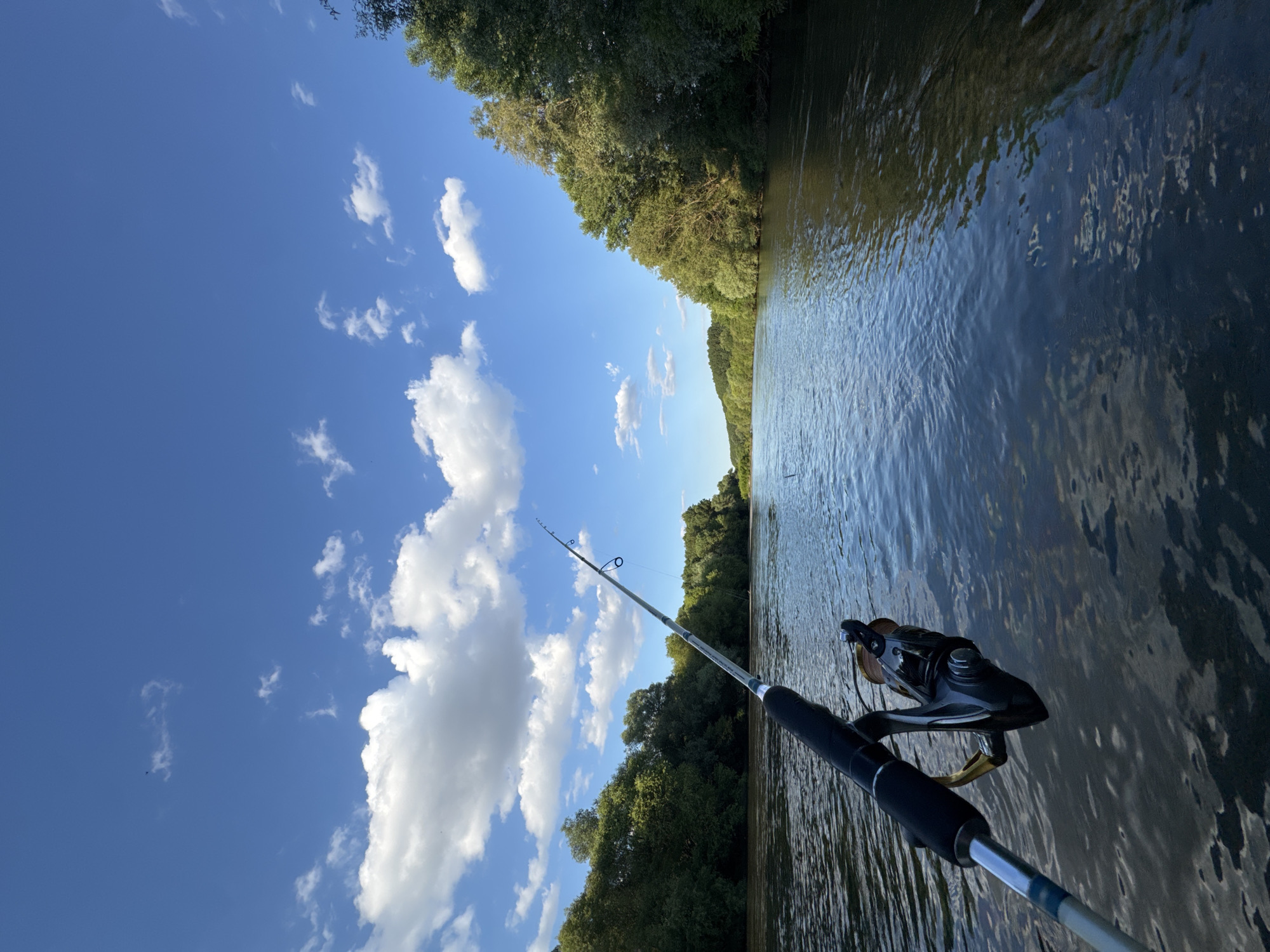 A fishing rod and reel are held by an unseen person, pointed towards a flowing river. The riverbanks are lined with lush green trees. The sky above is a vibrant blue, dotted with fluffy white clouds. The sun is shining, indicating it is daytime. The water appears calm, with ripples on its surface reflecting the surrounding trees and sky. The image evokes a sense of peace and tranquility, suggesting a leisurely fishing trip in a natural setting. The time of day appears to be late morning or early afternoon, given the bright sunlight and the position of the shadows. The weather seems pleasant, with clear skies and moderate cloud cover. The location is likely a river in a wooded area, with no specific landmarks visible to identify the exact spot. There is no visible text in the image.