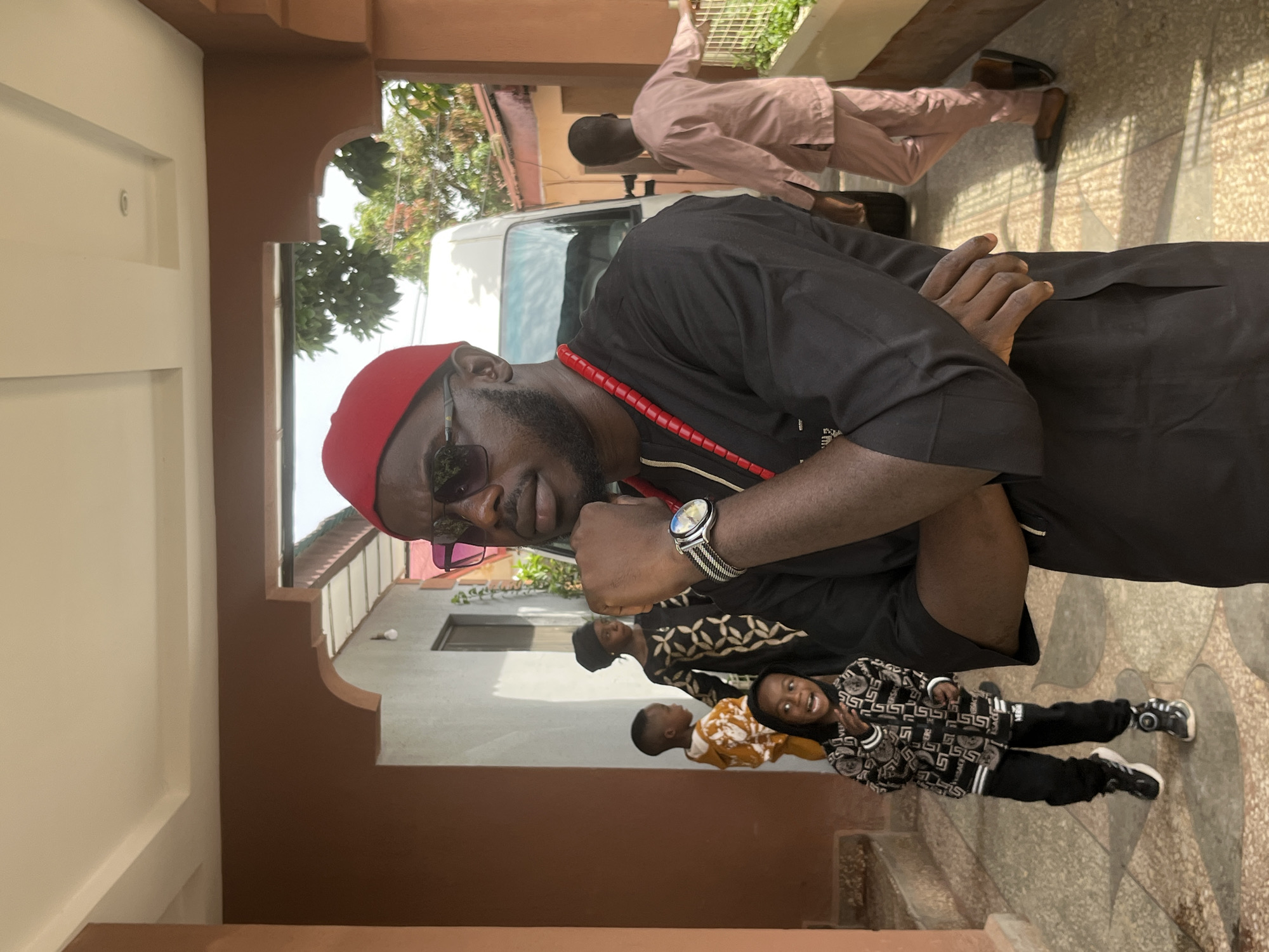 This image captures a scene in Jos, Nigeria, featuring a group of people in an outdoor, covered walkway. The central subject is a man wearing a black traditional outfit, a red cap, sunglasses, and a red beaded necklace. He is standing with his arms crossed and his chin resting on his hand, looking towards the right of the frame.
In the background, several children are visible. One child in the foreground is wearing a black and white patterned jacket and pants, with a wide smile. Another child, to the left, is wearing a yellow hooded outfit. Further back, a young person in a black and white patterned dress and headscarf is also present.
To the right of the frame, a person in a light pink traditional outfit is seen walking away from the camera. Behind the central subject, a white van is parked. The environment appears to be a residential or institutional setting with tiled flooring and brown-painted walls and pillars. Sunlight casts shadows on the ground, indicating it is daytime.