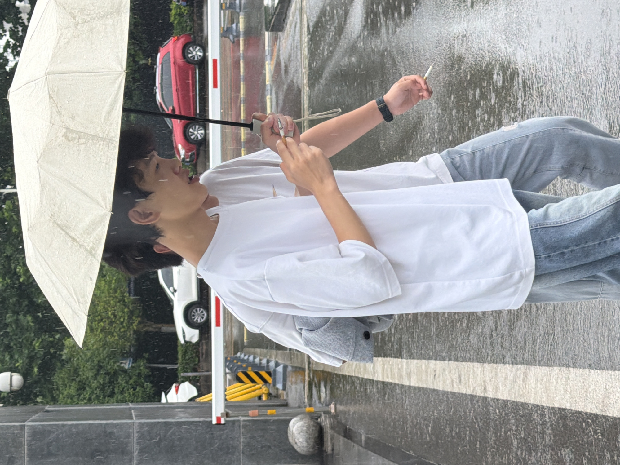 The image displays two individuals walking on a wet, dark asphalt surface during daylight rainfall. The foreground figure, a male wearing a white short-sleeved t-shirt and light blue denim jeans, holds a silver smartphone in his right hand. A grey garment is draped over his left arm. The second individual, partially obscured behind the first, also a male, wears a white t-shirt and light blue jeans. His right hand grasps the black shaft of a beige umbrella, while his left hand holds a lit cigarette; a dark wristwatch is present on his right wrist. In the background, a white dome-shaped security camera is visible on the upper left. A white traffic barrier with a red stripe extends horizontally across the midground, fronting yellow and black striped bollards and dark concrete blocks. Beyond this barrier, multiple vehicles including a red SUV and a white SUV are visible on a street lined with green foliage. The wet ground exhibits reflections and a prominent white lane marking. This scene is located in the city of Jiangjin, China.