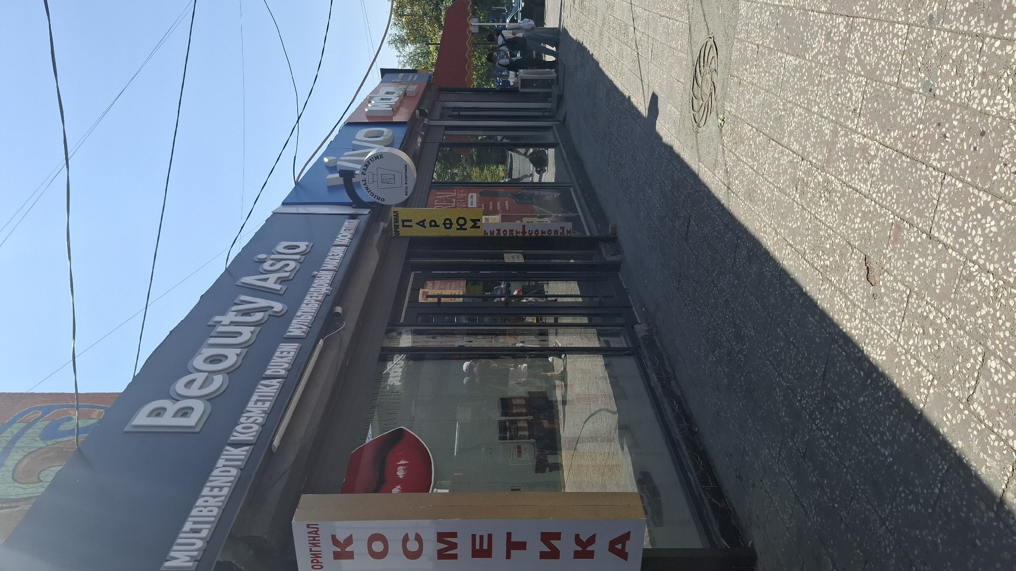The image shows the exterior of a commercial establishment in Shymkent, Kazakhstan, on a sunny day. The main sign above the storefront reads "Beauty Asia" in large, white, three-dimensional letters, with smaller text below it in Russian indicating "MULTIBRENDTIK KOSMETIKA DUKENI MUL'TIBRENDOVYY MAGAZIN KOSMETIKI." To the left of the main sign, a vertical sign with red letters on a yellow background reads "КОСМЕТИКА." The storefront features large glass windows and doors, reflecting the sky and surrounding buildings. Inside the window, various cosmetic products and displays are visible.
Further to the right, there are additional storefronts with signs. One sign, circular with a stylized perfume bottle, reads "ORIGINAL PARFUME" and "REAL PERFUMES." Next to it, a vertical yellow sign has red Cyrillic text that appears to be advertising "ПАРФЮМ" (perfume) and "РЕМОНТ" (repair). Further down the block, partially obscured signs for "VIVO" and "MOON" are visible.
On the sidewalk in front of the shops, several people are present. Two individuals, one wearing a white cap and t-shirt, and another wearing a dark t-shirt and shorts, are standing near the entrance of one of the shops. Further to the right, two individuals, one wearing a white t-shirt and jeans and the other in a dark t-shirt and jeans, are standing near what appears to be a counter or display. A car is partially visible behind them. The sidewalk is paved with speckled concrete tiles. A circular manhole cover is visible on the sidewalk in the right side of the frame. The scene is illuminated by sunlight, casting shadows across the pavement.