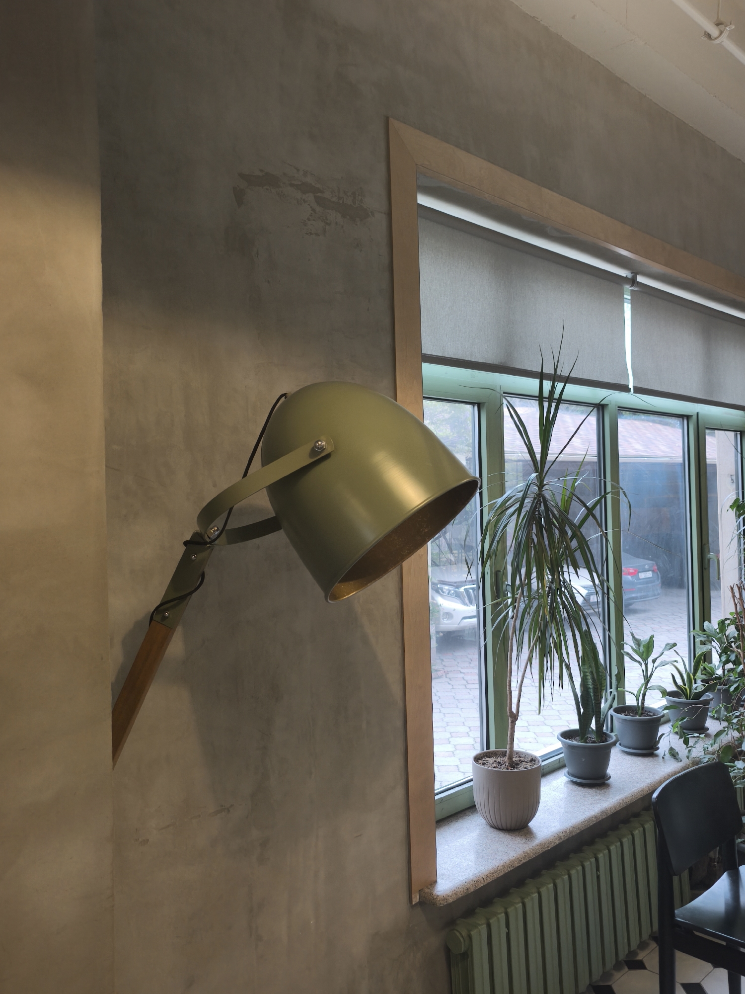 The image shows the interior of a room with a focus on a wall, window, and various decorative elements. A green metal floor lamp with a wooden base is positioned against a textured, gray wall. The window has a light wood frame and green muntins, with a partially drawn roller shade. A row of potted plants sits on the windowsill, including a Dracaena Marginata, a snake plant, and several other plants with broad leaves. Outside the window, the view shows a paved area with cars parked, suggesting a residential or commercial setting. The room appears to be well-lit with natural light. A portion of a black chair is visible in the lower right corner, and a green radiator is beneath the windowsill. The floor is a black and white patterned tile.