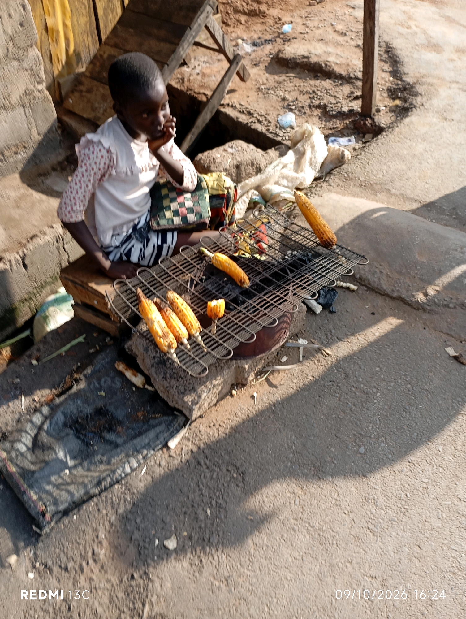 A young child sits near a makeshift grill made of stones and metal, roasting corn on the cob. The child wears a white shirt with pink polka dots and a patterned skirt. Several corn cobs are visible on the grill, some charred from the heat. The scene appears to be outdoors, possibly in a market or street setting, with dirt ground and a concrete path. In the background, there are some wooden structures and what looks like trash. A date and time stamp, "09/10/2026 16:24", is visible in the bottom right corner, along with the text "REDMI 13C".
