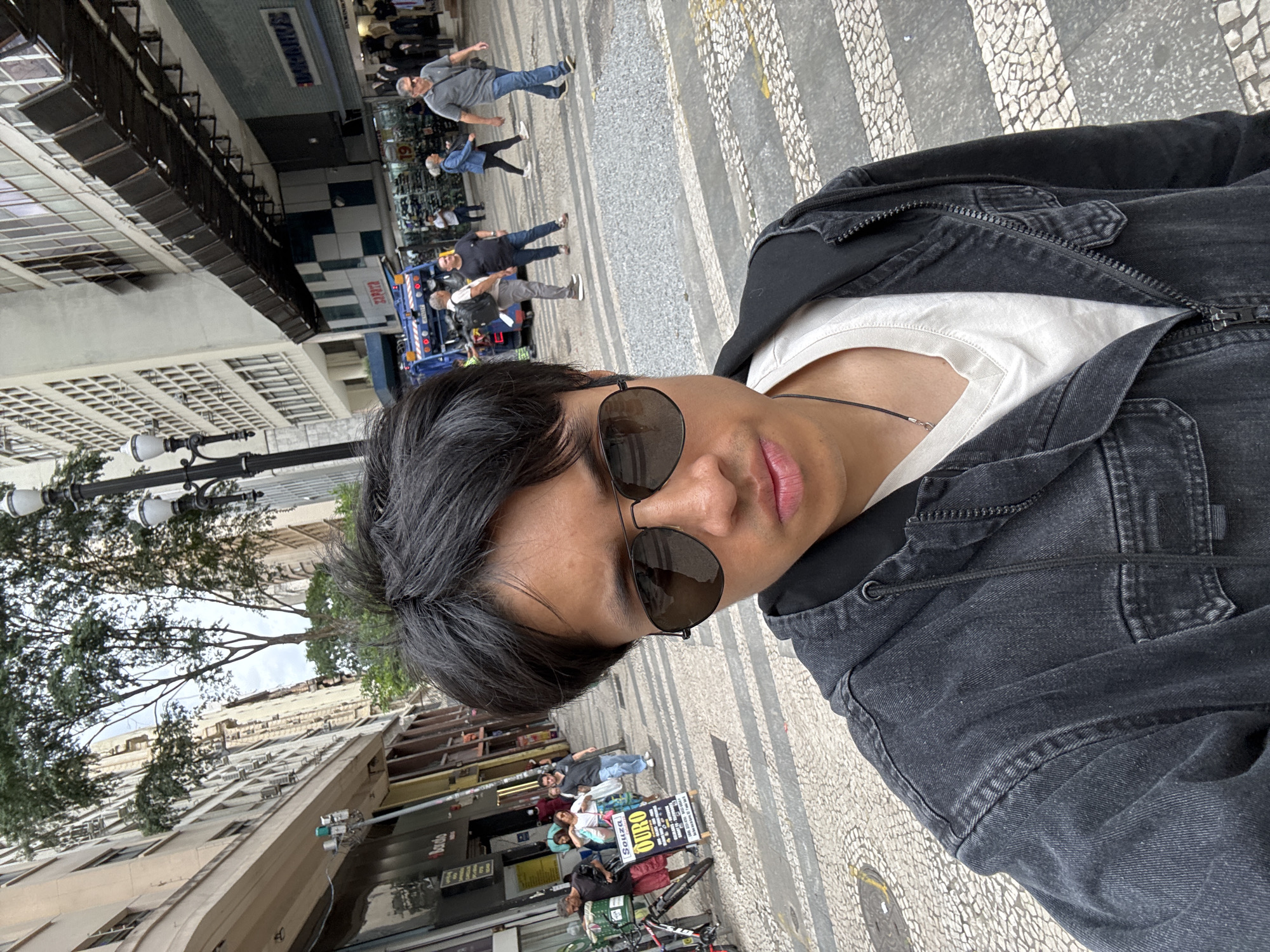 The image shows a selfie taken on a street in São Paulo, Brazil. A person with dark hair and sunglasses is facing the camera, wearing a black denim jacket over a white t-shirt. Behind them, a busy street scene unfolds. Pedestrians are walking on a pavement with a distinctive black and white pattern. Street vendors are positioned near buildings. In the background, there is a partial view of buildings with a mix of architectural styles and a blue maintenance vehicle with yellow lights.