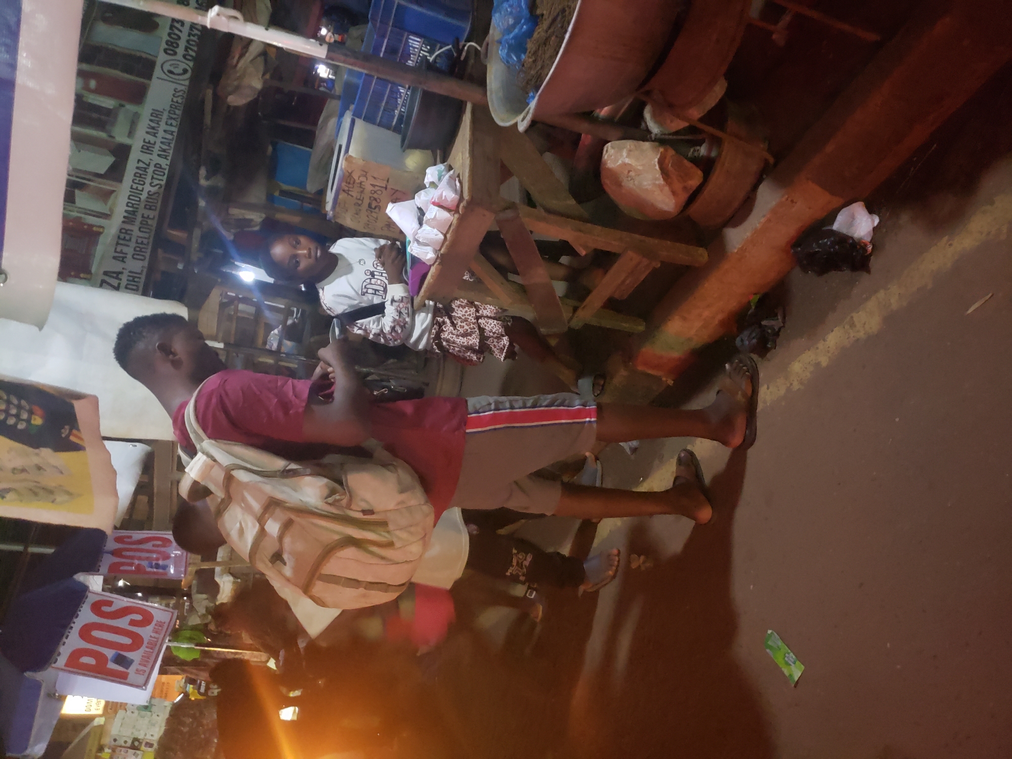 The image captures a bustling night market scene in Ibadan, Nigeria. In the foreground, a young man wearing a maroon shirt, grey shorts, and a light-colored backpack walks or stands with his bare feet visible, facing towards the right side of the frame. Behind him, a young woman in a light-colored top with a "Dior" logo is seated behind a wooden stall, looking towards the viewer.
The market features various wooden structures and stalls. A large reddish-brown basin filled with dark material sits on a wooden stand. Small white packages are visible on the woman's stall. The ground is dark, possibly asphalt, and contains some litter, including a green wrapper and a black plastic bag.
The scene is brightly lit by artificial light sources, creating strong glares and deep shadows, suggesting it is evening or night.
Visible text on signs provides commercial and location details: "ZA, AFTER MARDIEGRAZ, IRE AKARI, DHL, ORELOPE BUS STOP, AKALA EXPRESS" along with phone numbers "080738" and "0703716." Other signs declare "POS IS AVAILABLE HERE" and display "ALEX ARENAJU 2102958811," likely contact information for a vendor. The overall atmosphere is that of an active, informal street market.