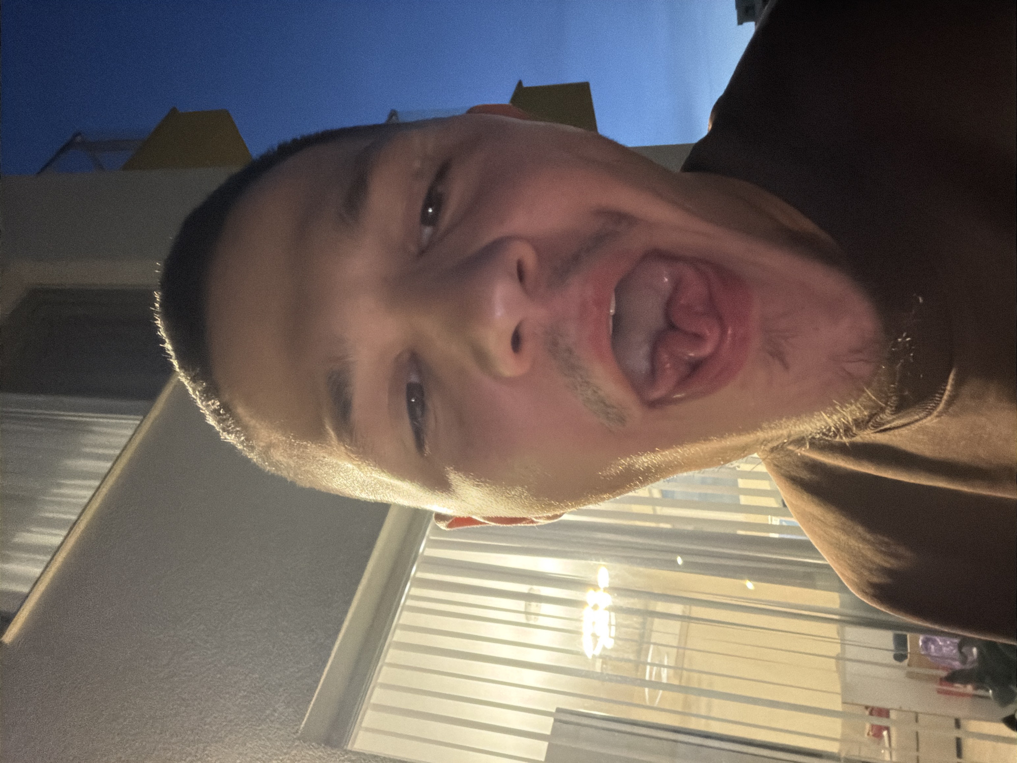 A close-up, selfie-style portrait shows a person with a shaved head and a light mustache and goatee. The person's mouth is open wide, with their tongue sticking out and curled. The background suggests an outdoor urban setting at dusk or night, with a building visible on the right and vertical blinds and interior lighting seen through a window on the left. The person is wearing a brown sweatshirt.