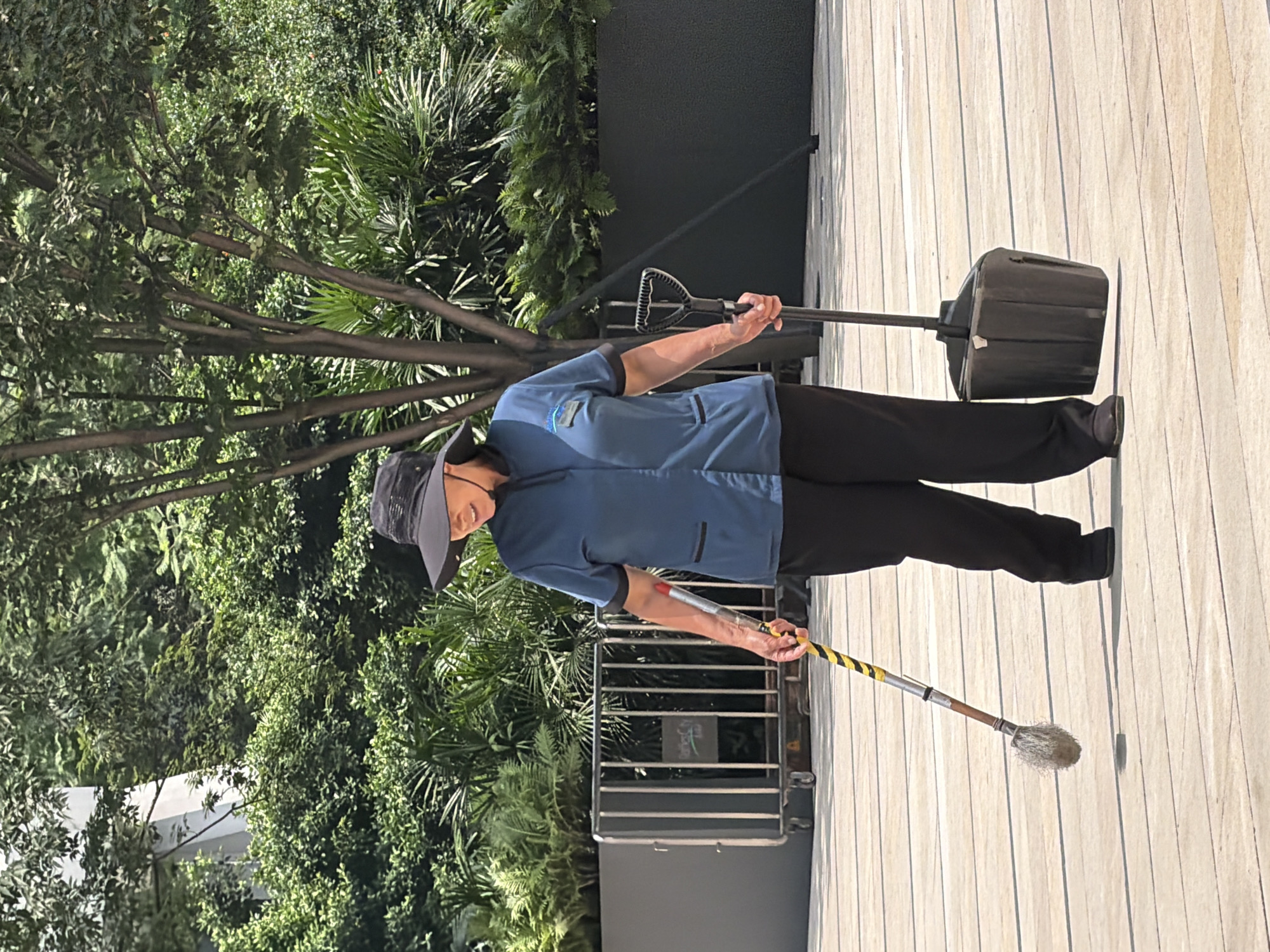 A person wearing a wide-brimmed hat and a blue uniform is holding a broom and a dustpan. The person is standing on a wooden boardwalk surrounded by lush greenery and trees. In the background, a metal fence with vertical bars is visible. The scene appears to be an outdoor urban environment, possibly a park or a plaza. The lighting suggests it is daytime. The person's attire indicates they are likely engaged in cleaning or maintenance work. The person is looking down and to the left, with their right hand holding the dustpan handle and their left hand holding the broom handle. The broom has a yellow and black striped shaft.