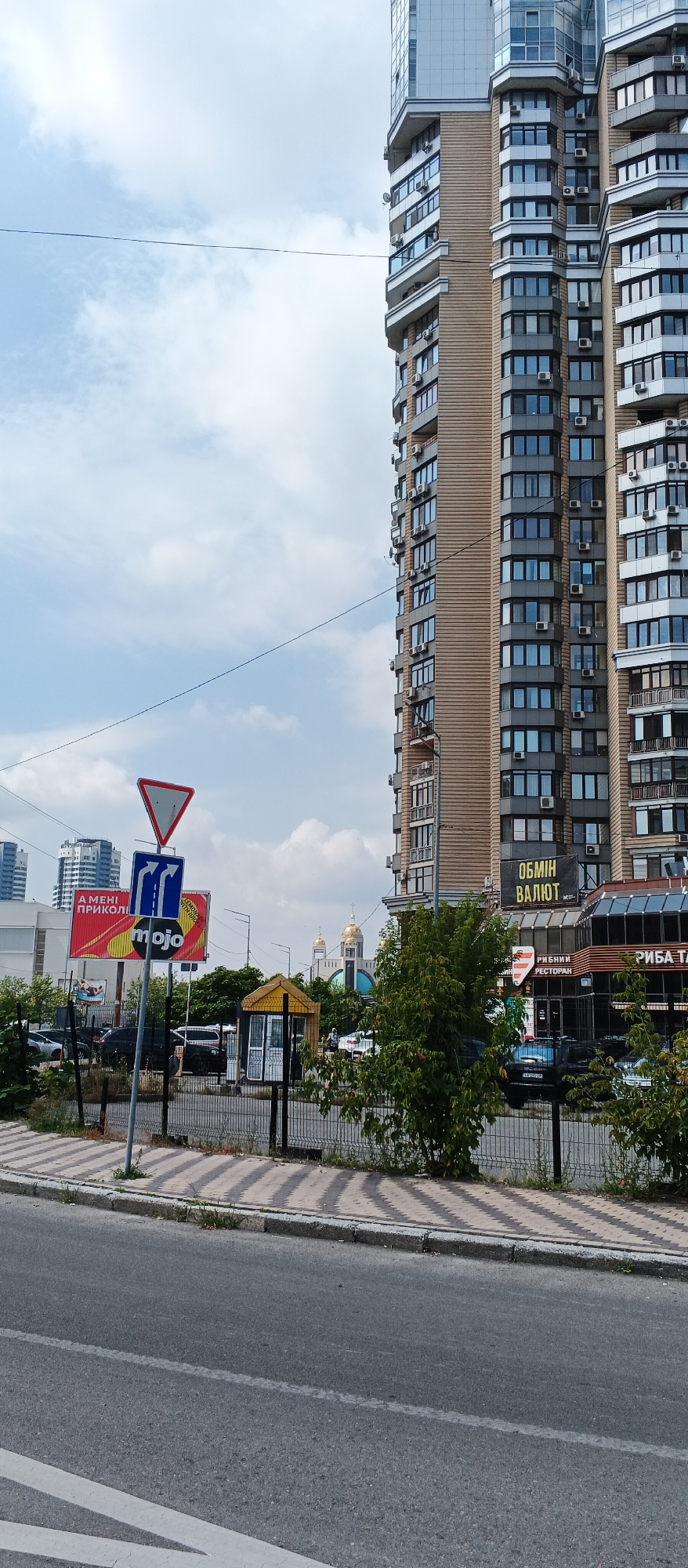 A street scene in Kyiv, Ukraine, captures a modern multi-story residential building with beige and gray facades, featuring numerous balconies and windows. In the foreground, a paved road with white lane markings runs alongside a sidewalk with a patterned surface. A metal fence separates the sidewalk from a grassy area with trees and bushes. Several traffic signs are visible, including a yield sign, a directional sign with arrows indicating lanes, and a red sign with Ukrainian text. A small yellow structure, possibly a kiosk or bus stop, stands near the fence. In the background, two golden-domed churches are visible, suggesting a religious or historical area. Several cars are parked along the street. The sky is partly cloudy.