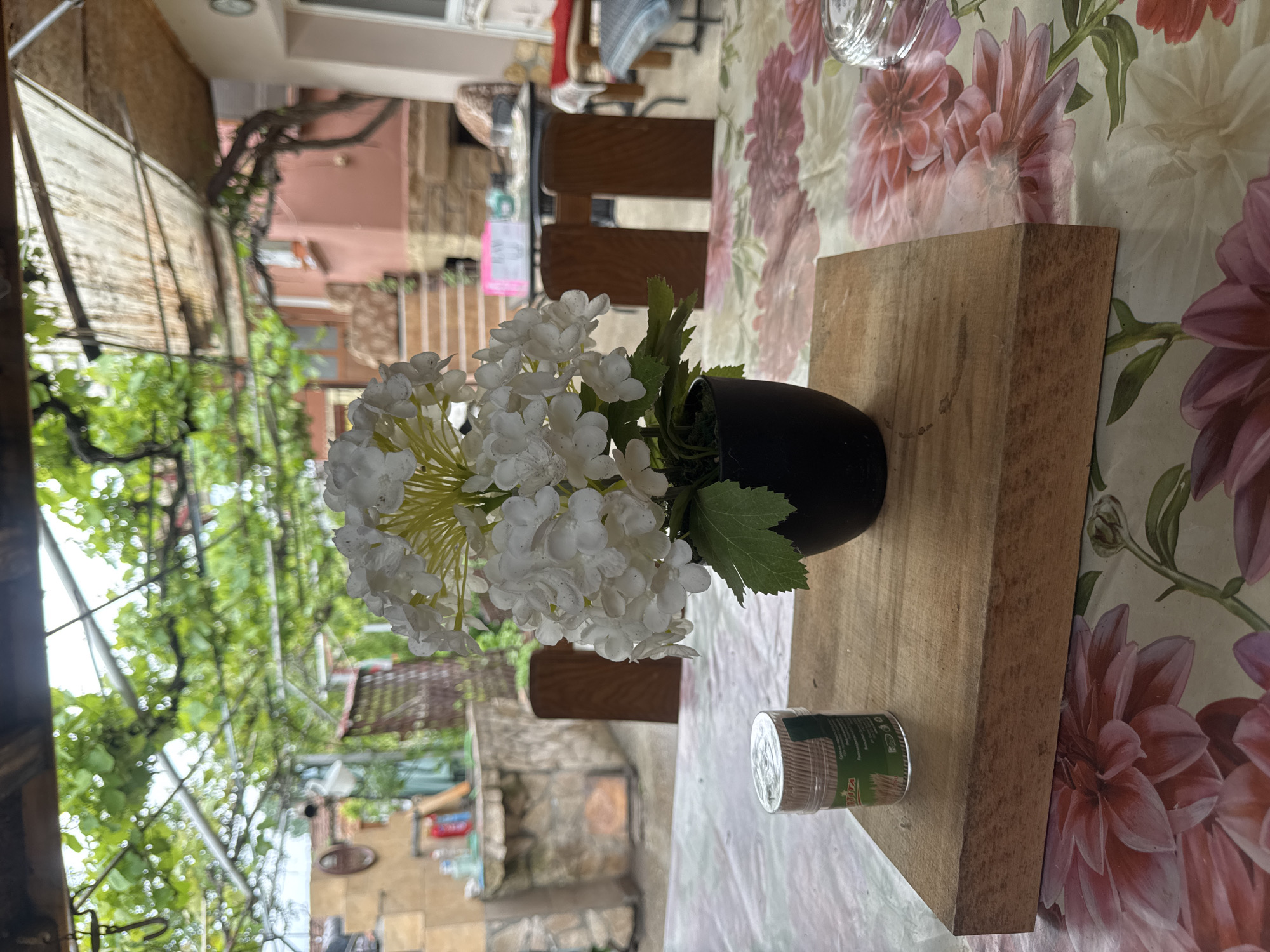 The photograph shows a still life arrangement on a table, likely outdoors in Lehchevo, Bulgaria. The table is covered with a floral-patterned tablecloth and holds a wooden cutting board. On the board, there is a black vase containing artificial white flowers with green leaves, and a cylindrical container of toothpicks. The background shows a view of the town with buildings, greenery, and a pergola. The overall scene suggests a casual, outdoor setting, possibly a patio or balcony in a residential area.