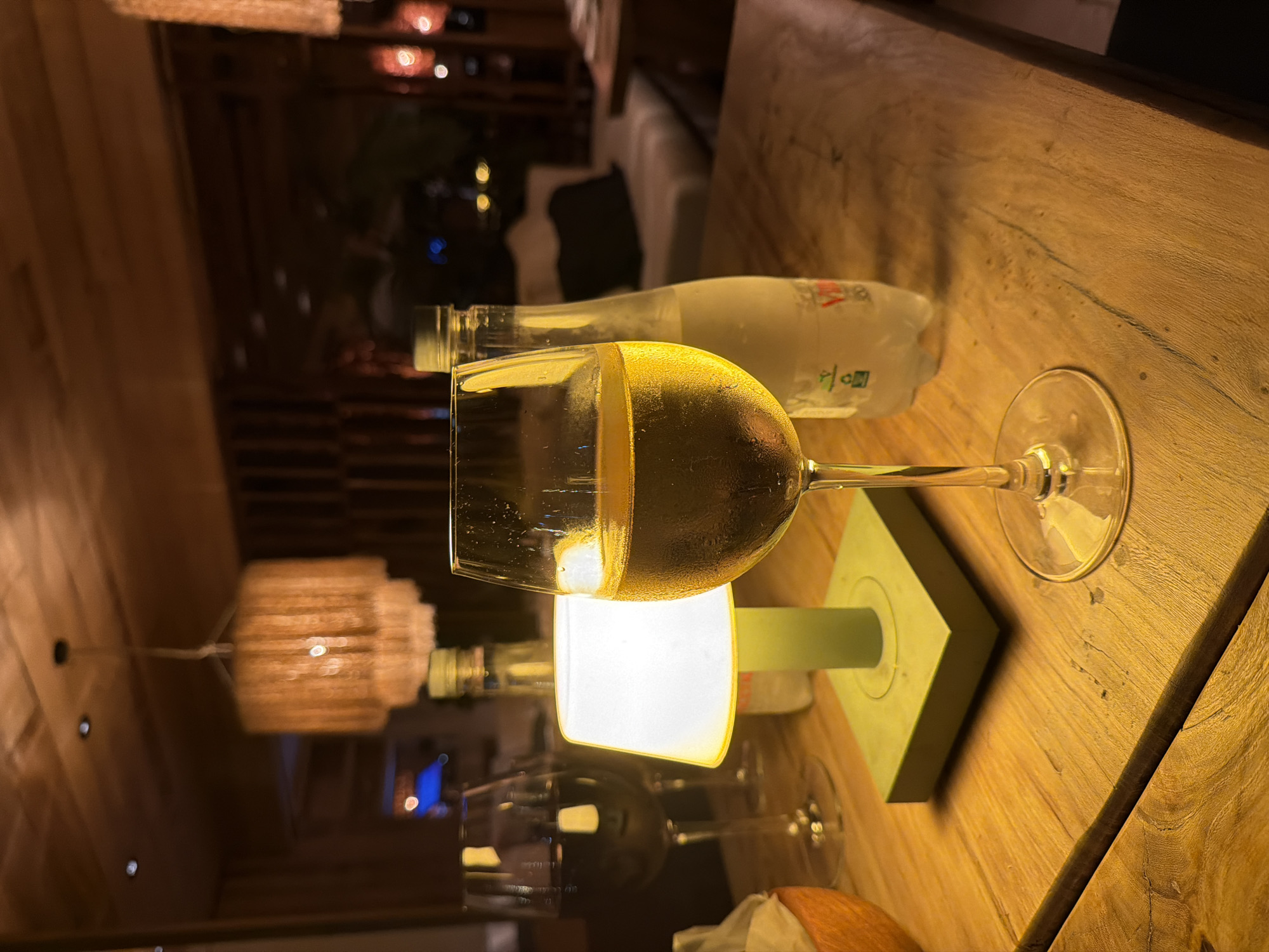 The image shows a close-up of a wine glass filled with a light-colored liquid, likely white wine. The glass is positioned on a wooden table next to a bottle of water. A small, illuminated lamp with a square base and cylindrical shade casts light onto the scene. In the background, other wine glasses are visible on the table, along with a blurry view of the restaurant interior, including a decorative woven light fixture. The image appears to be taken in a restaurant with a warm, ambient lighting scheme. While the image does not provide specific information to confirm its location in Buenos Aires, Argentina, the elements suggest a modern, upscale dining establishment.