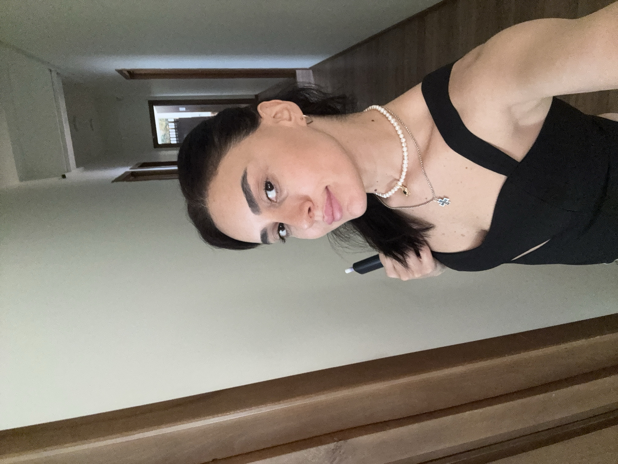 A woman with dark hair and brown eyes is taking a selfie in a hallway. She is wearing a black top and pearl necklaces. She is holding a black electronic cigarette. The hallway has light-colored walls and wooden doors. There is a glimpse of a room at the end of the hallway, which appears to be furnished. The lighting is natural and soft, suggesting it may be daytime. The overall scene is domestic.