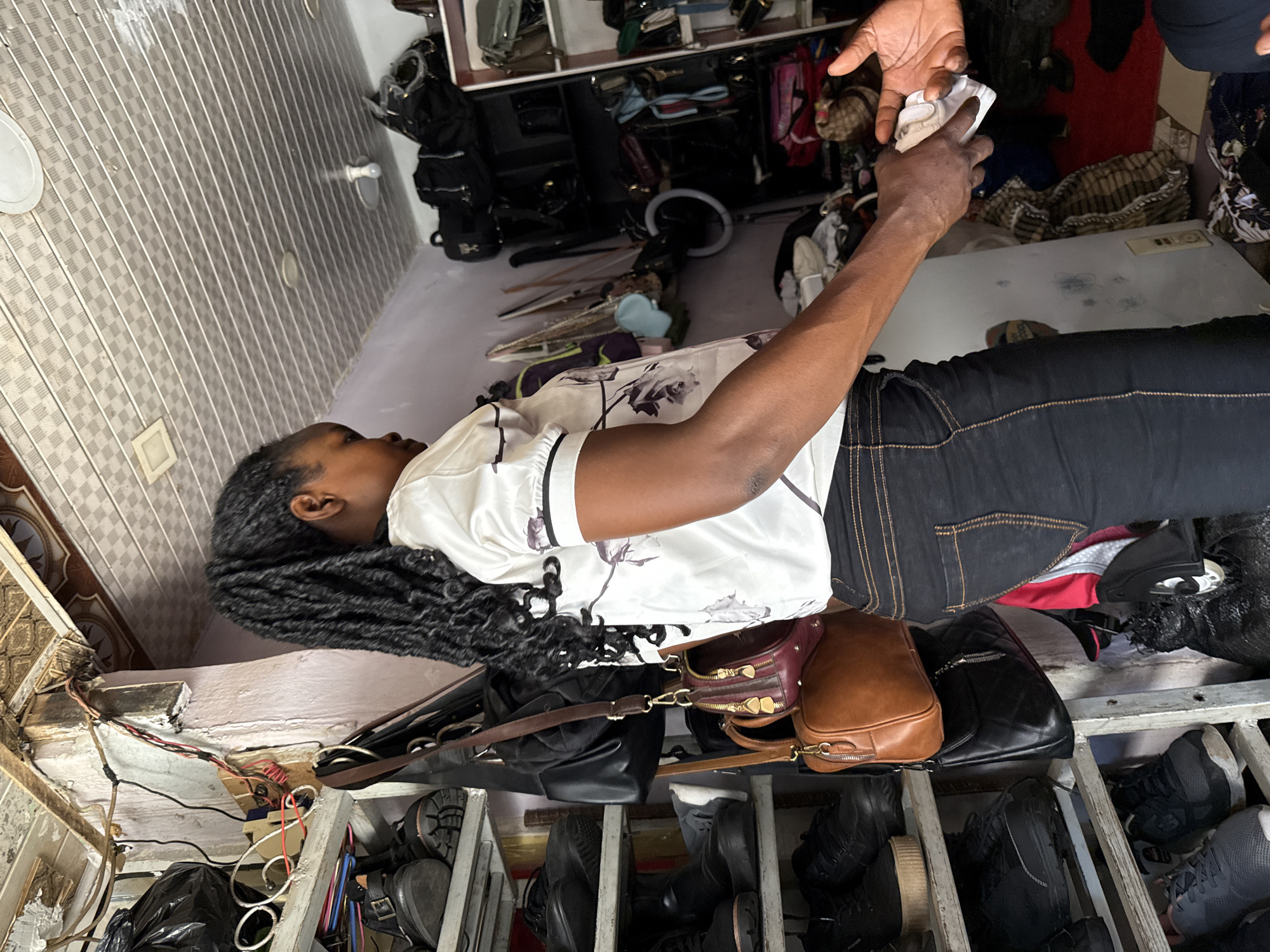 A dark-skinned woman with braided hair stands in a cluttered shop in Abuja, Nigeria. She is wearing a white shirt with a floral pattern and dark jeans. She is reaching out to another person, whose hand is visible, to receive a white object, possibly money. The shop is filled with shelves stacked with shoes and bags. The ceiling is made of checkered tiles, and wires are visible along the wall. The overall impression is of a busy, informal retail environment.