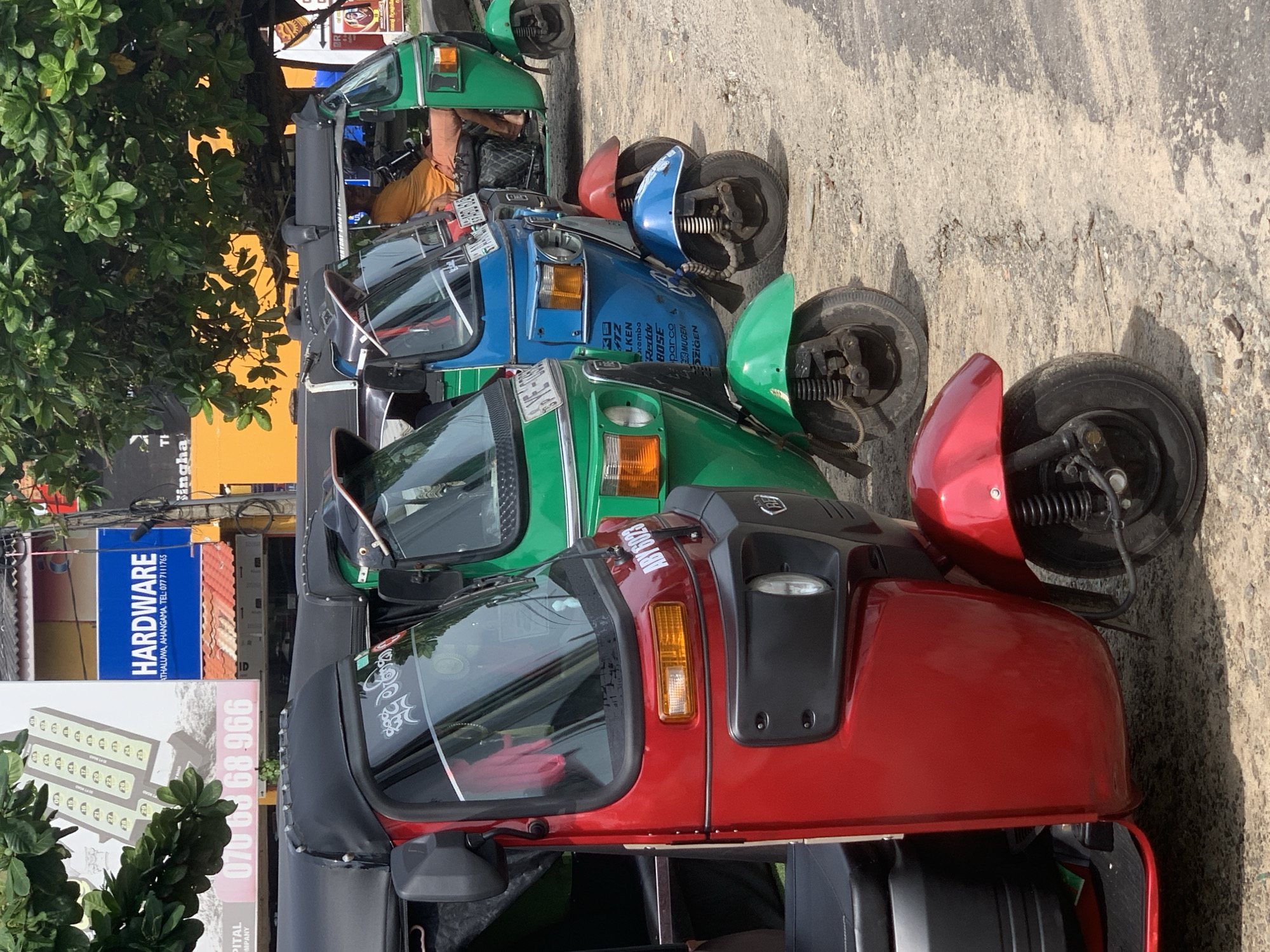 The image shows a row of four auto rickshaws, also known as tuk-tuks, parked on a street. The tuk-tuks are in different colors: red, green, blue, and another green. A man is sitting inside the first green tuk-tuk. The tuk-tuks are closely parked, and the front of each vehicle is visible.
A sign reading "Hardware" and "Kathaluwa, Ahangama" indicates the location is likely in Kathaluwa, Sri Lanka. There is a phone number and additional commercial signage visible nearby. The street surface appears to be asphalt or concrete. Green foliage is visible in the foreground, adjacent to the business signs.