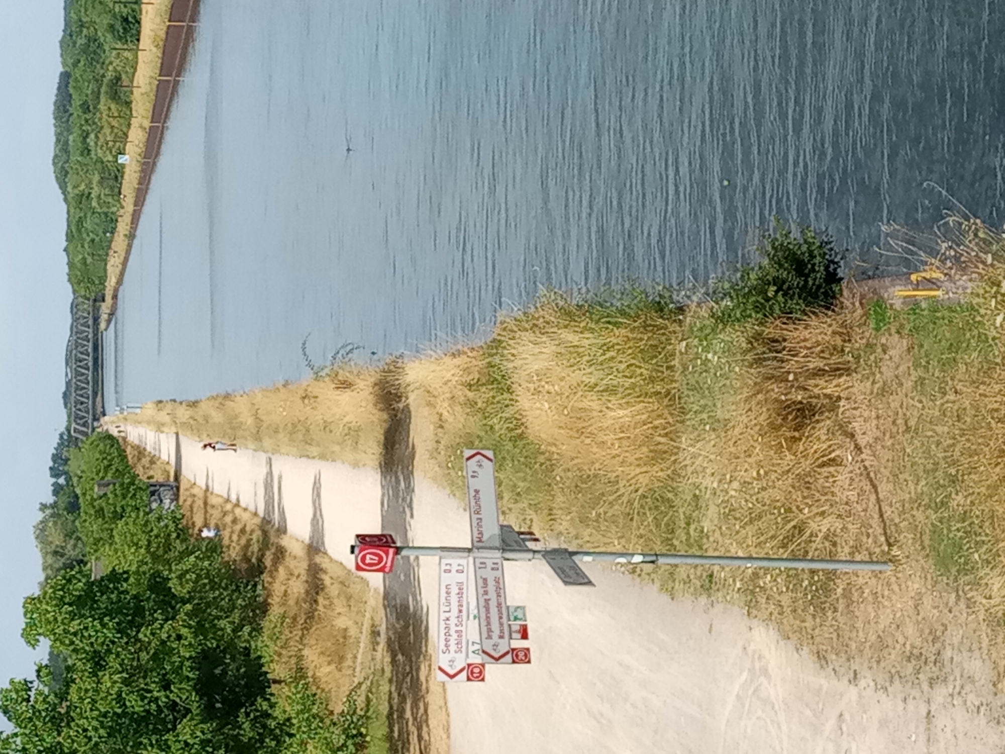 The image shows a canal with a path running alongside it. A signpost stands on the path, pointing in different directions. The signpost has a red and white marker with the number 17. Several signs with place names and distances are attached to the post, including "Seepark Lünen 0.3", "Schloß Schwansbell 0.7", "Marina Rünthe 9.8", "A7", "Bergwerksiedlung 'An Luran' 1.8", and "Wasserwanderastplatz 0.8". A person is walking on the path in the distance, and another person is sitting on the side of the path. The canal is wide and calm, with trees and vegetation on the banks. A railway bridge can be seen in the background. The scene appears to be in Lünen, Germany, given the presence of "Seepark Lünen" on one of the signs.