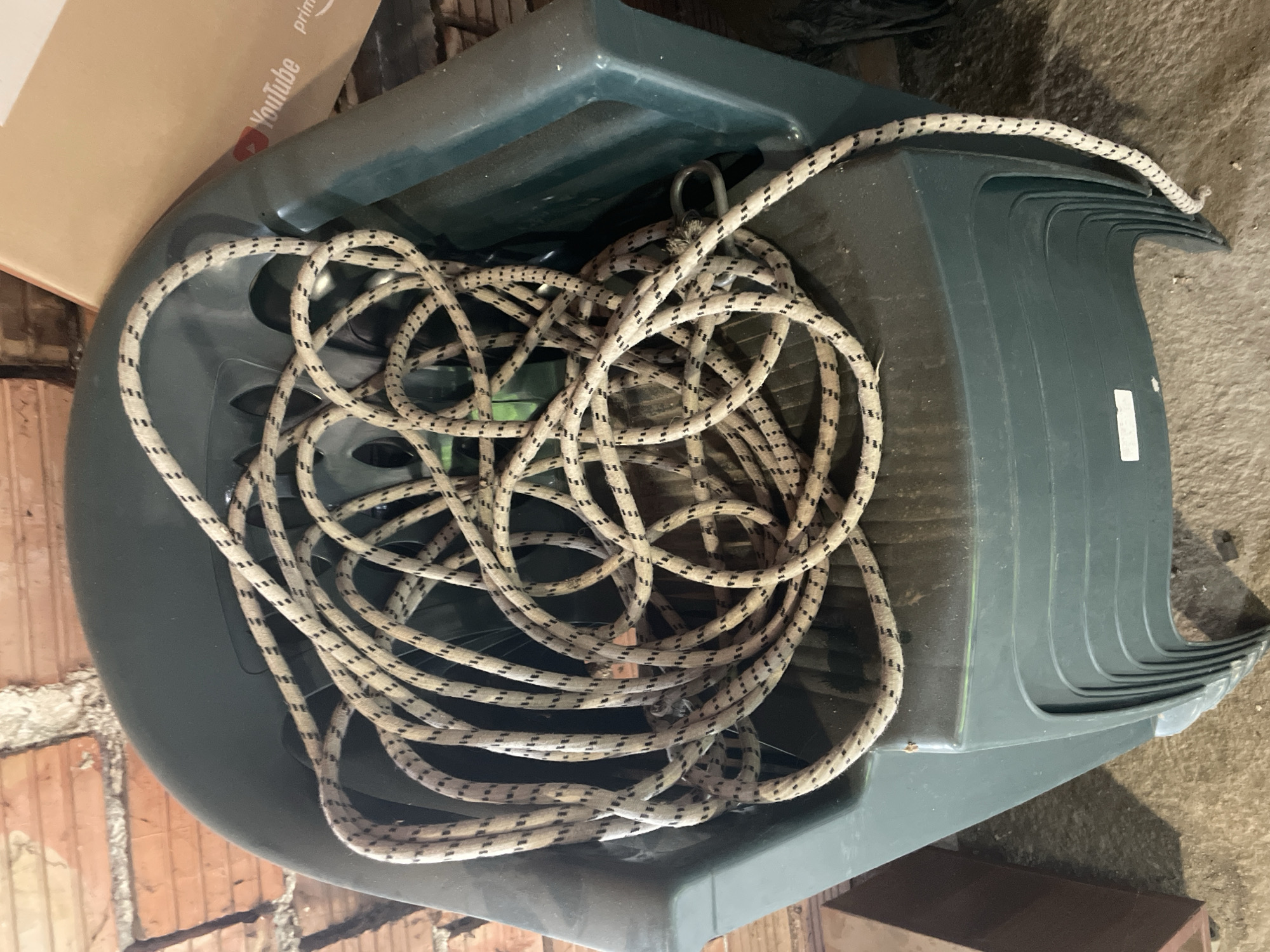 The image displays a dark green, likely plastic, sled or toboggan, viewed from an overhead perspective. A thick, beige and black speckled rope is coiled loosely inside the sled. A metal loop or hook, to which the rope is attached, is visible within the sled's interior.
The setting appears to be an indoor storage area, possibly a garage, basement, or shed. A brick wall is visible along the left side of the frame, and a concrete or dirt floor can be seen on the right and bottom. In the upper left background, part of a cardboard box is visible, featuring the "YouTube" logo and the word "prime."
There are no people present and no specific activity is taking place; the sled and rope appear to be stored. The lighting is somewhat dim, characteristic of an indoor storage space. The scene is situated in Ihtiman, Bulgaria.