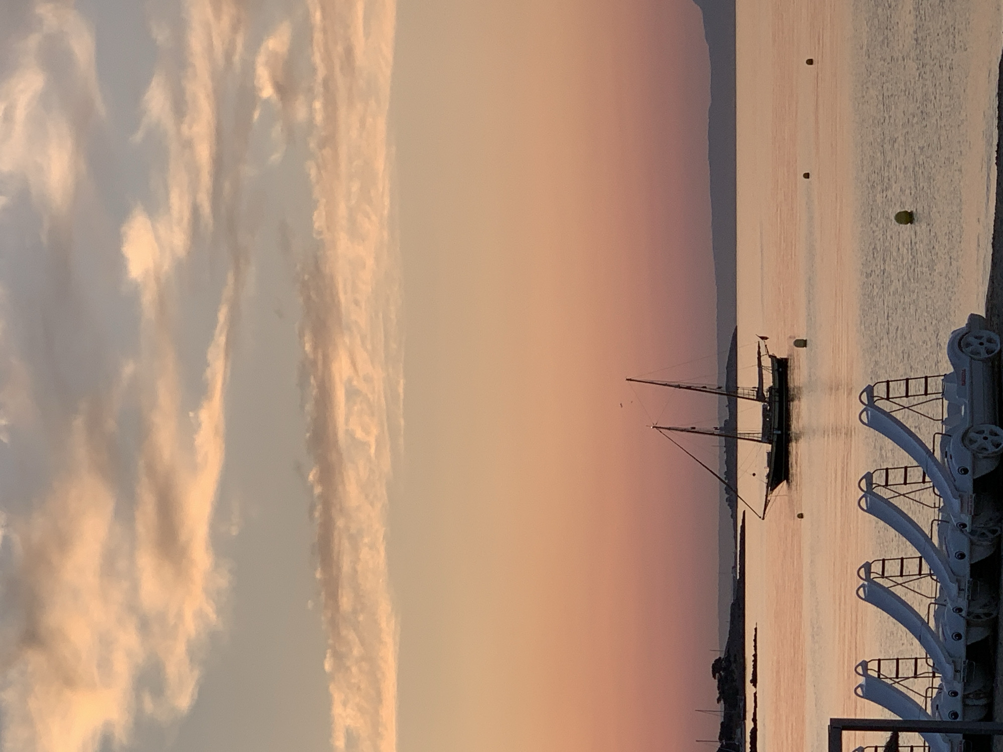 The image depicts a serene coastal scene at Palmanova, Spain, during what appears to be either sunrise or sunset. The sky is a gradient of soft oranges and pinks, with scattered wispy clouds catching the light. In the foreground, a row of white paddle boats with attached slides are lined up on the sandy shore, ready for use. Beyond them, the calm, reflective water of the Mediterranean Sea stretches out. A traditional sailboat with two masts is anchored in the distance, adding a classic maritime element to the view. The coastline is visible as a low, dark silhouette against the horizon, with hints of vegetation and structures. The overall atmosphere is tranquil and picturesque.