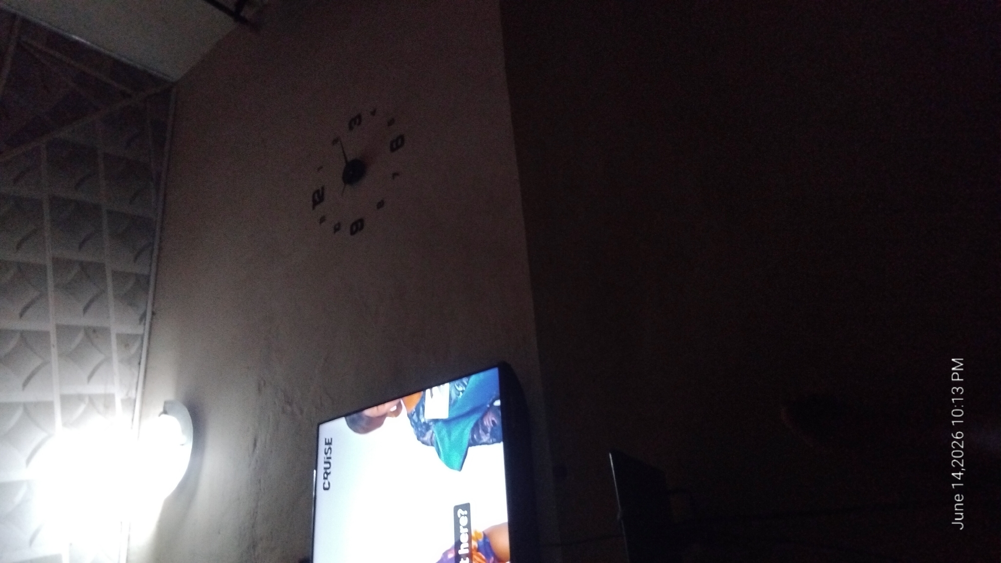 The image is a dimly lit interior shot. In the upper left, a fluorescent light fixture illuminates a textured ceiling with a diamond pattern. Below the ceiling, a portion of a television screen is visible, displaying the word "CRUISE" and an image of two people. To the right of the television, a black wall clock with Arabic numerals is mounted on a light-colored wall. The clock hands indicate a time close to 10:15. The lower portion of the image is dark, with text in the bottom left corner reading "June 14, 2026 10:13 PM". There is no indication of the specific city or location.
