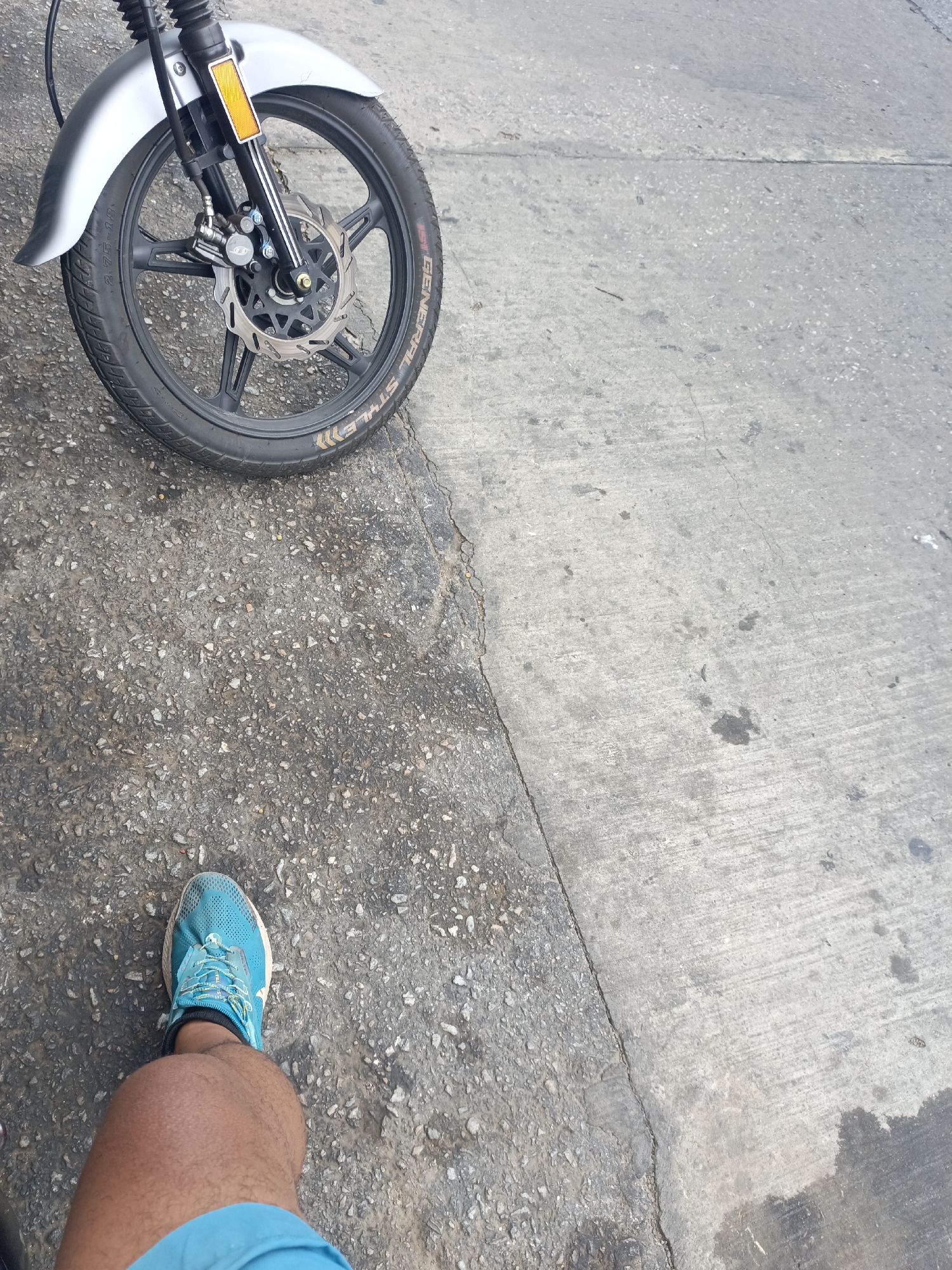 The image depicts a partial view of a motorcycle front wheel parked on a two-toned concrete surface, likely a street or sidewalk. A person's leg and foot, clad in a blue running shoe and matching shorts, are visible in the lower left corner. The surface consists of a rough, pebbly concrete section alongside a smoother, more uniform concrete slab. The scene is in an urban environment. There is no specific information indicating the location is Juan Griego, Venezuela.