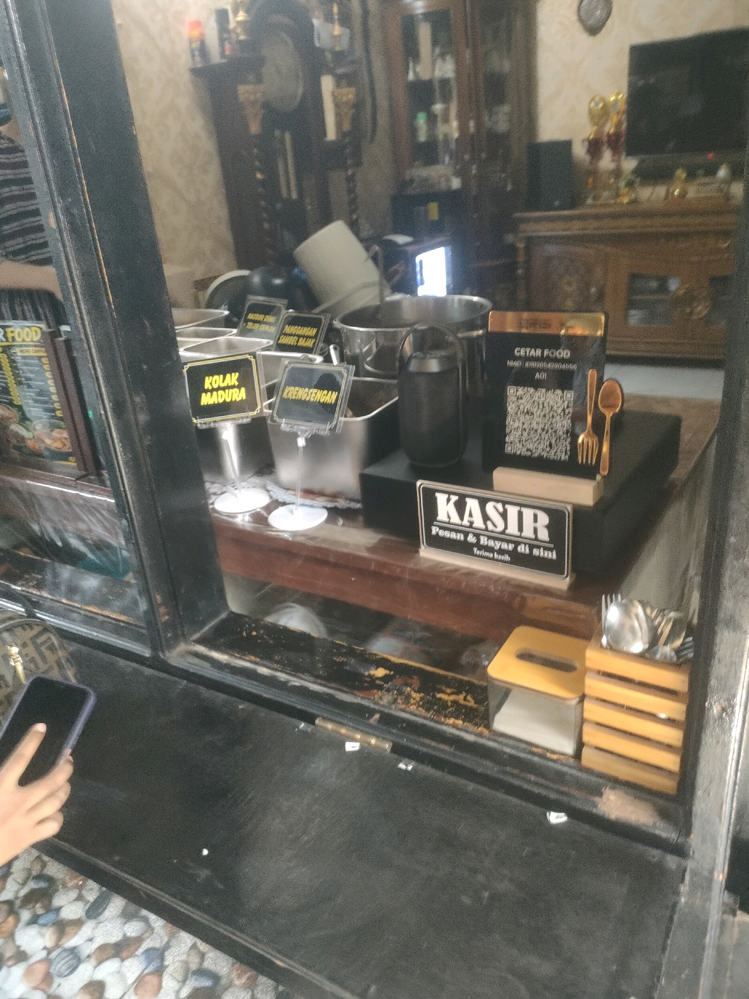 A hand holds a smartphone in the foreground, with a portion of a glass window reflecting a restaurant interior. Inside the restaurant, various food items in metal containers are visible. Signs indicate "KOLAK MADURA" and "KRENGSENGAN." A "CETAR FOOD" sign with a QR code and cutlery is also present, along with a "KASIR" sign that reads "Pesan & Bayar di sini Terima kasih." The background shows shelves with items, a television, and wooden furniture. The scene is likely a food stall or restaurant in Surabaya, Indonesia, given the presence of Indonesian language signs and common food items.