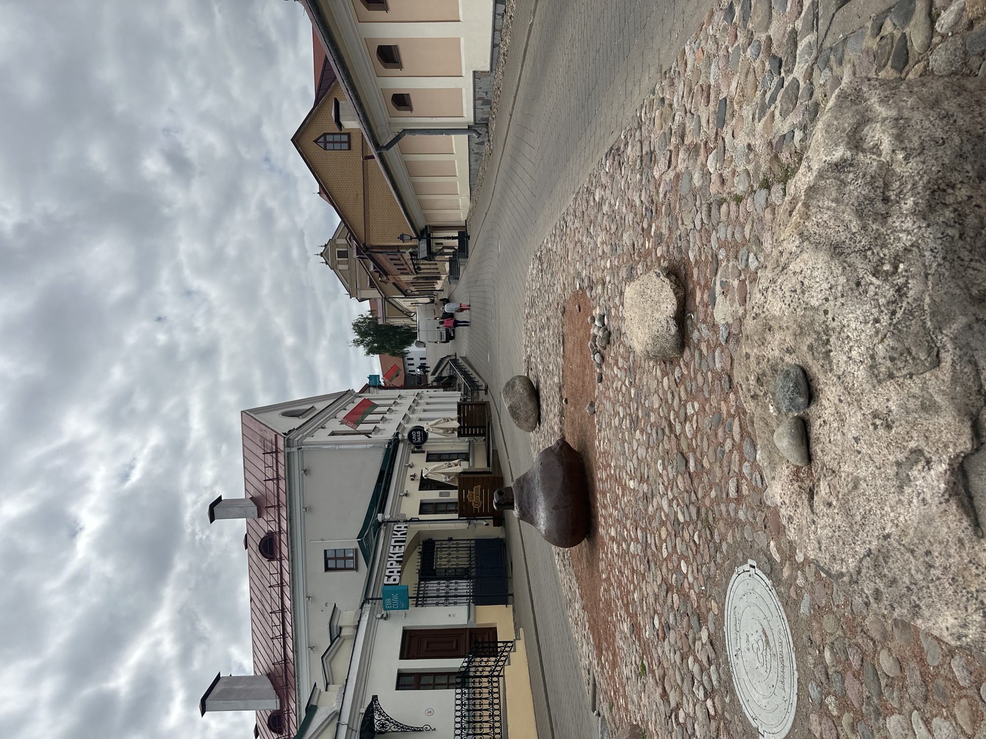 The image shows a cobblestone street in Minsk, Belarus. In the foreground, there is a large, dark, bell-shaped sculpture with a hole in the top, placed on a circular bed of red gravel and smaller cobblestones. Several large rocks are also scattered around the sculpture. A circular manhole cover is visible in the lower left of the frame.
The street curves upwards and to the right, lined with buildings on both sides. On the left, a white building with a red roof features several shops, including one with a sign in Cyrillic that appears to say "БАРКЕПКА" and another with "ELVA CLINIC". A Belarusian flag is visible hanging from one of the buildings. On the right, the buildings have yellow and cream-colored facades with red roofs.
In the background, two people are walking up the street. The sky is overcast with gray clouds. The overall impression is of a historic urban streetscape.