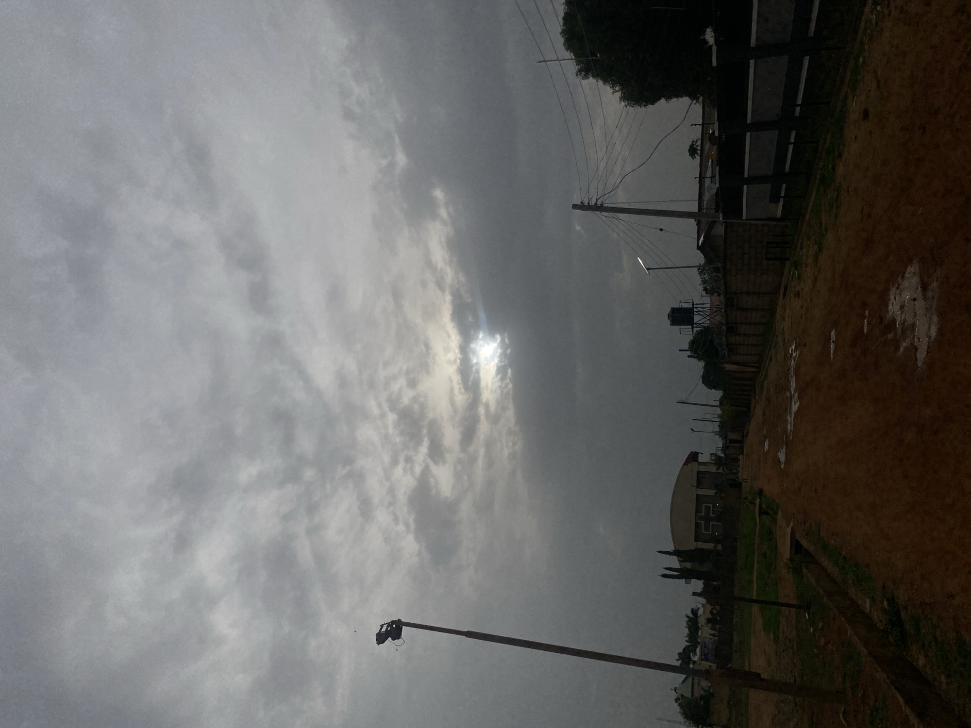 The image shows a landscape in Barden Yana, Nigeria, under a cloudy sky with a glimpse of the sun. There's an unpaved road or path, possibly wet. Along the path are buildings, power lines, and poles. A building with a cross on it is visible in the distance.