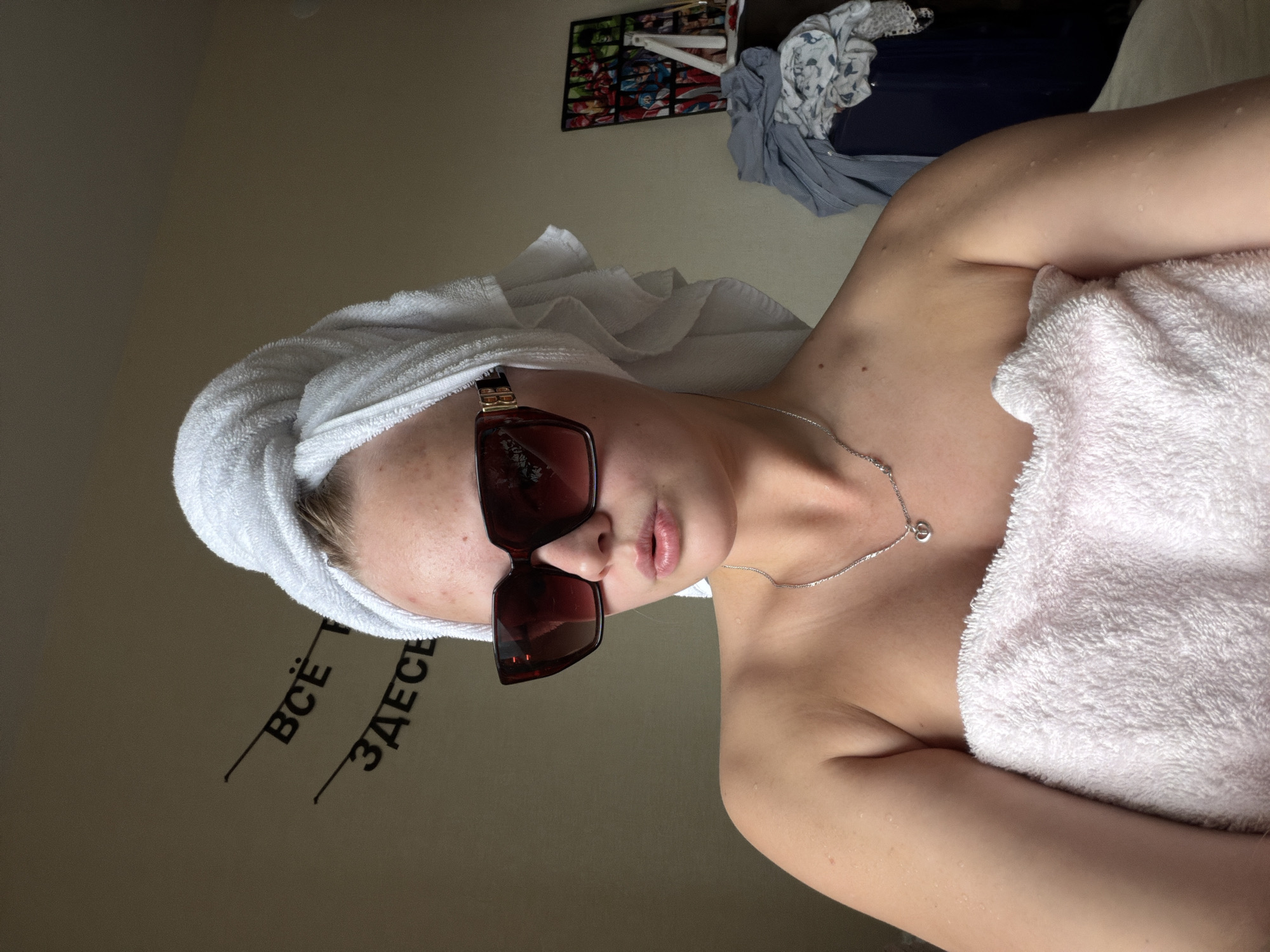 The image shows a young woman captured in what appears to be a casual, post-shower or self-care moment. She is positioned diagonally across the frame, with the original image being rotated 90 degrees clockwise. The woman has a white towel wrapped around her head like a turban and another large white towel wrapped around her body, covering her chest and shoulders. She wears large, dark-lensed sunglasses with thick, dark brown frames, obscuring her eyes. Her lips are slightly parted. A thin silver chain necklace with a small, circular pendant is visible around her neck, resting on her collarbone. Her exposed skin, particularly on her shoulder and arm, appears dewy or wet with small water droplets.
In the background, there is a plain, light brownish-grey wall. On this wall, two words in black Cyrillic script are visible, each underlined: "ВСЁ" (VSE), meaning "EVERYTHING," and "ЗДЕСЬ" (ZDES'), meaning "HERE." Above and to the right of the woman's head, a framed picture or poster with multiple panels displaying what looks like comic book characters is affixed to the wall. Further to the right, a pile of clothes, including a light blue or grey shirt and darker garments, is casually placed, possibly in a basket or on a surface. The setting appears to be an indoor, private space, consistent with a home environment in Izhevsk, Russia, given the visible Russian text. The lighting suggests an indoor setting with diffused light.