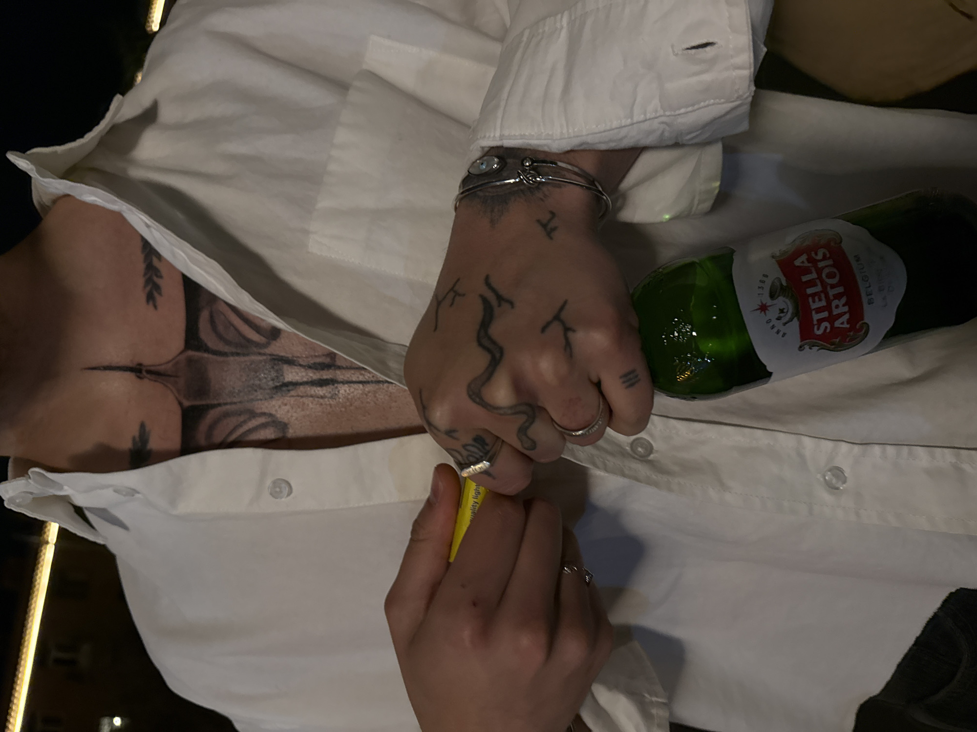 The image shows a person's torso wearing a white, button-down shirt, partially unbuttoned to reveal a chest tattoo. The person has tattoos on their hands and is wearing rings and a bracelet. They are holding a yellow object in one hand and a green glass bottle of Stella Artois beer is partially visible. It is not possible to identify Yerevan, Armenia as a location based solely on this image.