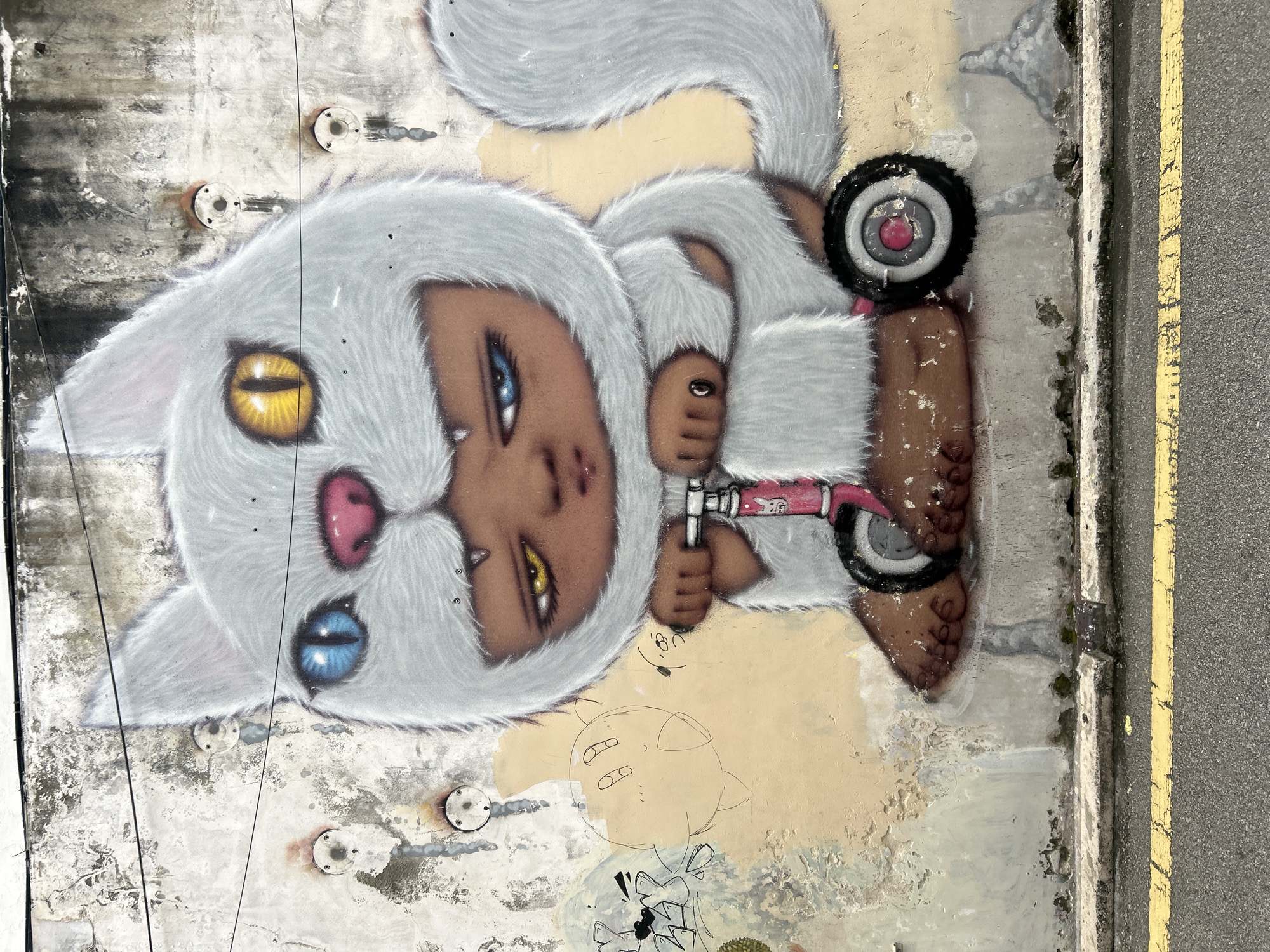 This image showcases a piece of street art on a weathered, pale yellow and grey wall in George Town, Malaysia. The artwork features a cartoonish, anthropomorphic cat character with a fluffy white costume, complete with ears and a tail. The character has distinctively mismatched eyes: one bright blue, the other yellow with a black pupil. It has a dark brown face with a pink nose and sharp teeth visible. The cat is depicted sitting on a pink scooter with black wheels, holding onto the handlebars with its hands. Its feet, which resemble human feet with toes, are planted on the ground.
The wall itself is textured and shows signs of age, with chipped paint and staining. In the background, faint outlines of other stylized figures and shapes can be seen, suggesting a larger mural or a collection of graffiti. Several metal fixtures, possibly for electrical conduits or plumbing, are mounted on the wall. The bottom of the image reveals a section of asphalt road with a cracked double yellow line, indicating its urban setting. The overall impression is one of vibrant and whimsical street art contrasting with a decaying urban surface.