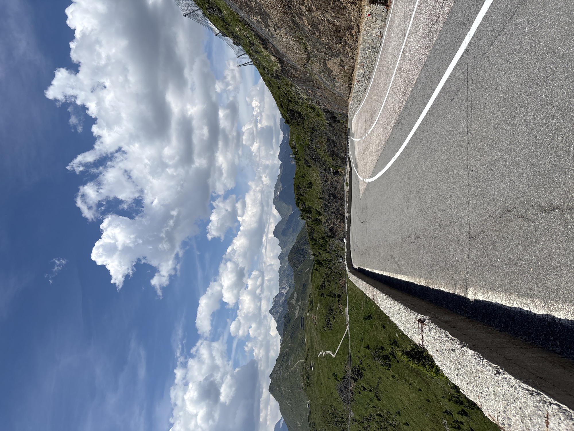 The image depicts a winding mountain road in the Italian Alps, likely near St. Leonhard in Passeier - San Leonardo in Passiria. The road curves sharply to the right, with a concrete barrier on the left and a steep, rocky embankment on the right. White lines mark the lanes of the asphalt road. Beyond the road, rolling green hills dotted with sparse trees stretch towards a majestic mountain range under a bright blue sky filled with white, puffy clouds. A mesh fence is visible on the embankment, designed to prevent rockfalls. The scene appears to be captured on a clear, sunny day. There are no people or vehicles visible in the image.