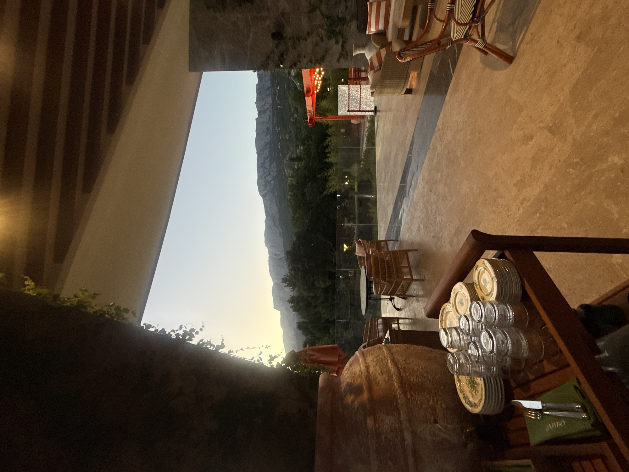 The image depicts an outdoor patio area of a restaurant or resort, overlooking a scenic mountain landscape at dusk. In the foreground, a large terracotta amphora stands prominently on the left, with a wooden cart laden with stacked plates and glassware to its right. A green napkin with silverware rests on the cart.
The patio floor is tiled with a light-colored stone. Several wicker chairs and tables are arranged on the patio, some with cushions. In the background, a transparent glass barrier separates the patio from a garden area. Beyond the garden, a red structure with string lights is visible, suggesting a separate seating or event space. The landscape transitions into a dense forest, with rugged mountains rising in the distance under a pale twilight sky. The location is identified as Симеизский поселковый совет, Ukraine. There are no people visible in the image.