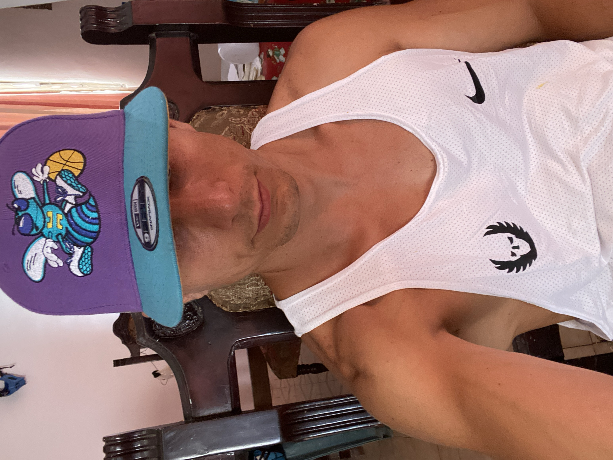 An indoor, eye-level portrait shot captures a man from the chest up, seemingly sitting or leaning against dark wooden furniture in Tinaquillo, Venezuela. The man has a neutral expression and is looking towards the camera. He wears a vibrant purple baseball cap with a light blue brim, featuring a cartoon character on the front, resembling a bee or insect holding a basketball, wearing sneakers, and displaying "1C" or "JC" on its chest. The hat's brim clearly shows "NEW ERA" and "SNAPBACK" text.
He is also wearing a white, mesh-like tank top. A black Nike swoosh logo is visible near his right shoulder, and another stylized black graphic, possibly a creature's head, is positioned on the lower left side of his chest. A small yellow stain is visible on the upper right side of his tank top. The background consists of dark, ornate wooden furniture, which appears to be part of a chair or sofa, and a light-colored wall. Reflections from an unseen light source are faintly visible in the background, especially in the darker areas of the furniture. The lighting suggests it is daytime.