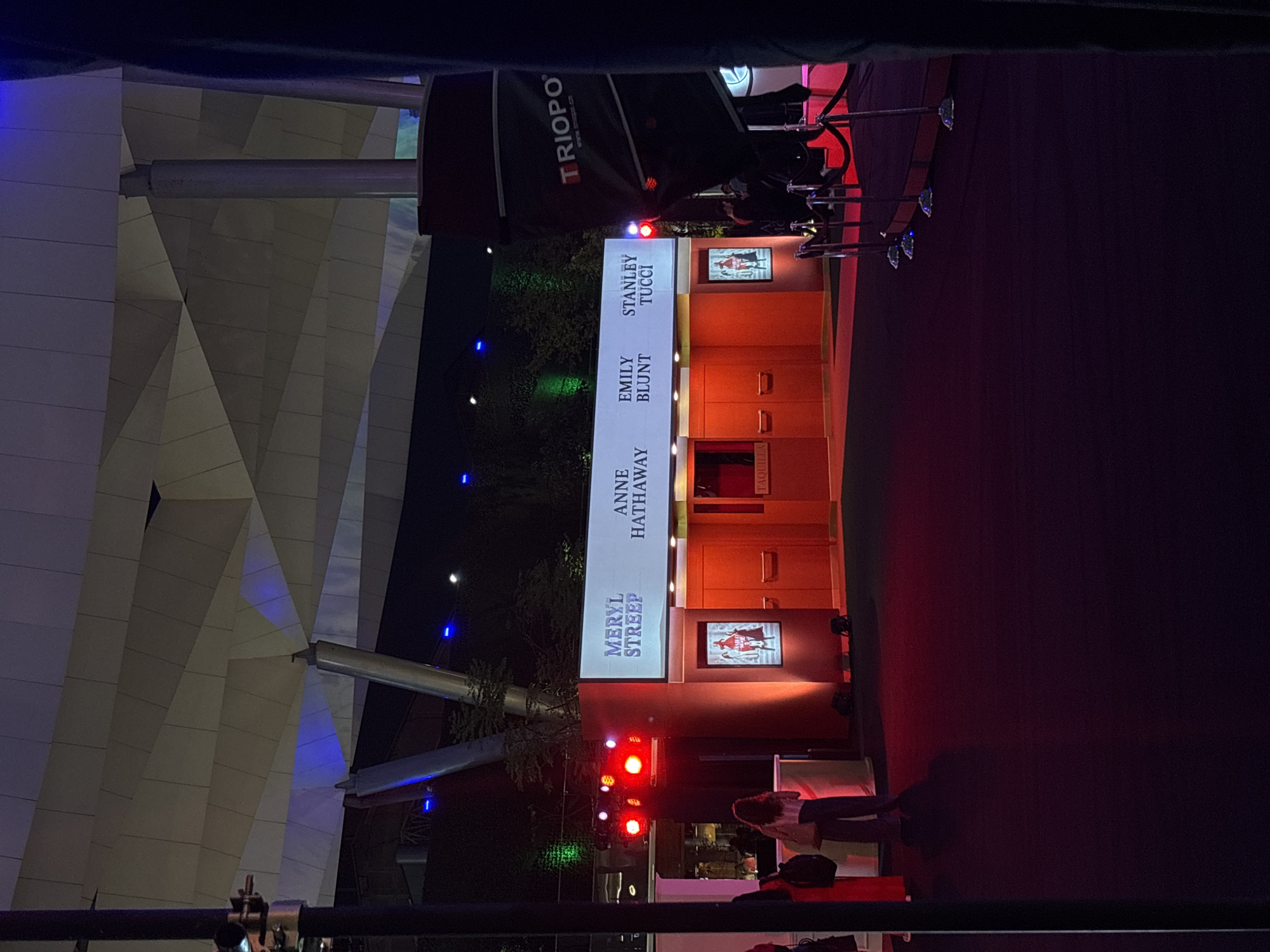 The image shows a red carpet event, likely a movie premiere, possibly at a location near Naucalpan de Juárez, Mexico. Visible on the backdrop are the names Meryl Streep, Anne Hathaway, Emily Blunt, and Stanley Tucci. There is a large structure behind the red carpet, appearing to be modern architectural design with triangular shapes and blue lighting. A film crew and camera are present, along with audience members and individuals moving around. The lighting includes red spotlights and more ambient lighting on the structure. The setup suggests a formal, public gathering, centered around the individuals named on the backdrop.