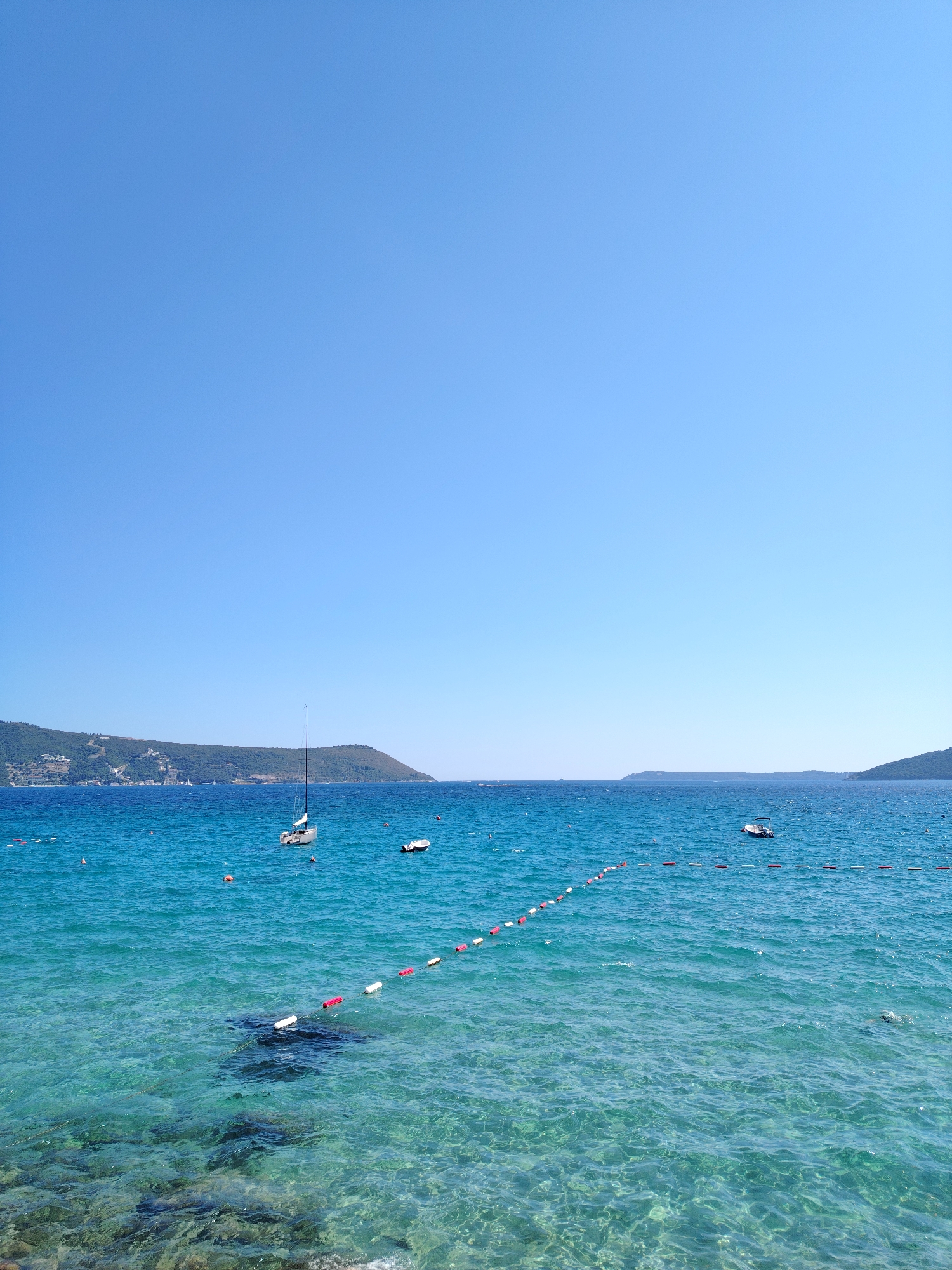 This is a horizontal, eye-level, medium shot of the Bay of Kotor in Herceg Novi, Montenegro, on a clear, sunny day. The sky is a cloudless, brilliant blue, taking up approximately the top half of the image. Below the sky, the turquoise waters of the bay are clear enough to see the rocky, submerged terrain beneath the surface in the foreground. A line of white and red buoys stretches diagonally across the water from the lower left towards the center-right of the image, demarcating a swimming area.
In the middle ground, a white sailboat with a tall mast is anchored. To the right of the sailboat, a small, white motorboat is also anchored. Further in the distance, along the left horizon, a green, hilly coastline is visible, dotted with buildings. The coastline extends across the horizon, with fainter hills visible in the far distance on the right. The water is calm, with gentle ripples reflecting the sunlight. A person is visible swimming on the far right of the image. The overall scene depicts a tranquil coastal environment.