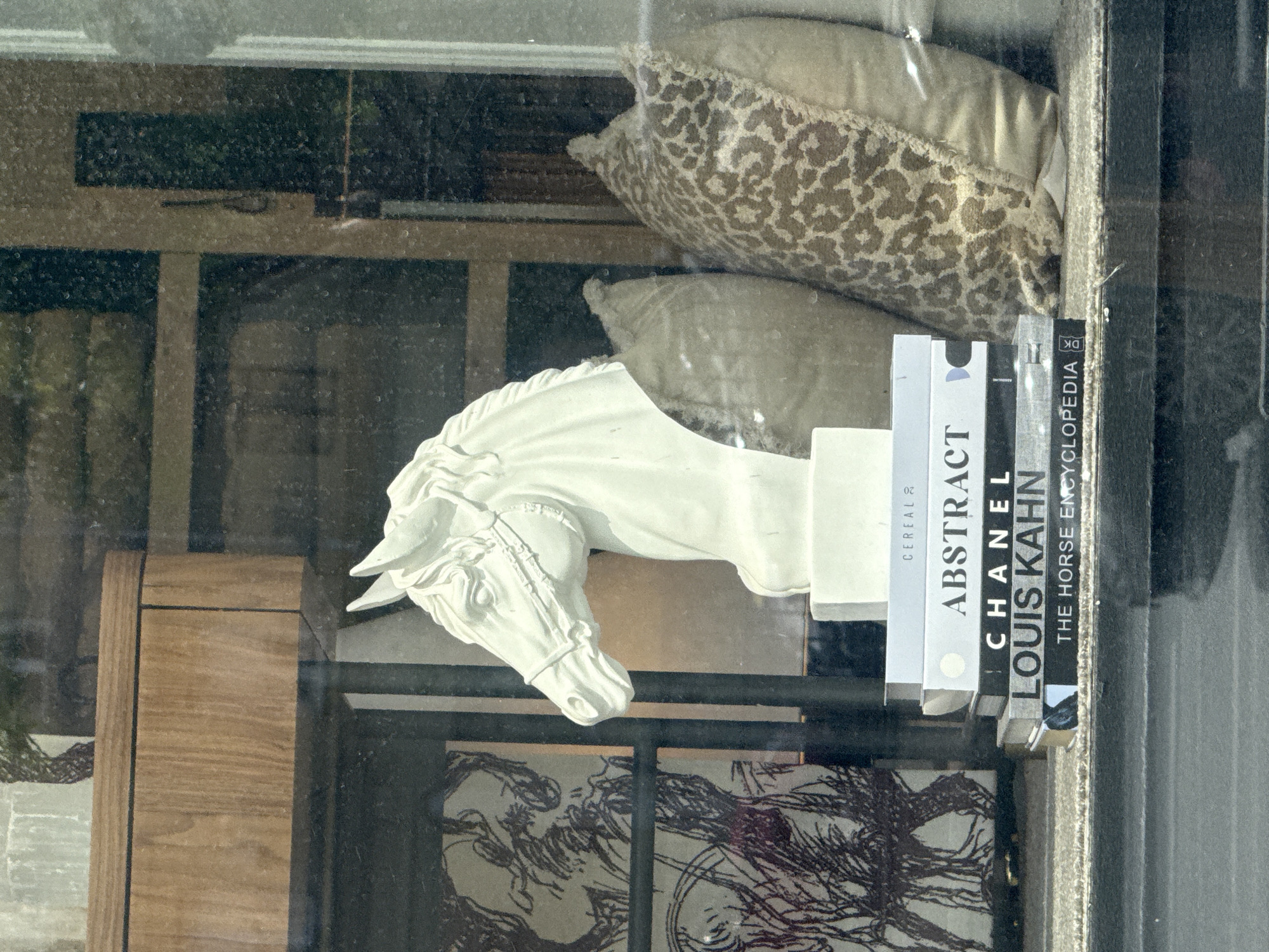 The image shows a storefront display. Dominating the view is a white, plaster horse head sculpture. Behind the sculpture are several books stacked horizontally, titles visible from left to right: "CEREAL", "ABSTRACT", "CHANEL", "LOUIS KAHN", and "THE HORSE ENCYCLOPEDIA". Above the books and sculpture are two pillows, one with an animal print. The display is behind a window, with reflections visible. The wooden framework of the window is also visible. Below the horse sculpture is another piece of artwork or decorative item featuring dark, sketched patterns on a light background. There is no indication of the location being in Moscow, Russia.