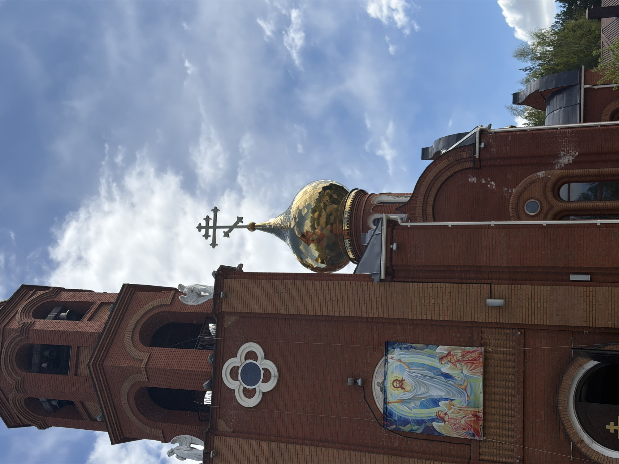 The image shows a tall, red brick Eastern Orthodox church in Chaikovsky, Russia, featuring a golden onion dome topped with a cross. The architectural details include arched windows in the bell tower, decorative white angel statues, a quatrefoil window, and a religious banner depicting Jesus with angels. The sky is blue with scattered white clouds.
