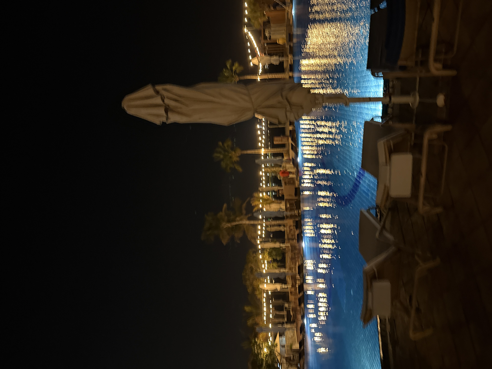 The image depicts a nighttime scene at a resort or hotel pool in Dubai, United Arab Emirates. The pool water is illuminated with reflections of string lights and palm trees that line the perimeter of the resort. Several lounge chairs are visible, some with cushions, suggesting they are ready for use. In the foreground, a large, closed umbrella stands near the pool. The overall atmosphere is serene and relaxing, typical of a resort setting at night.