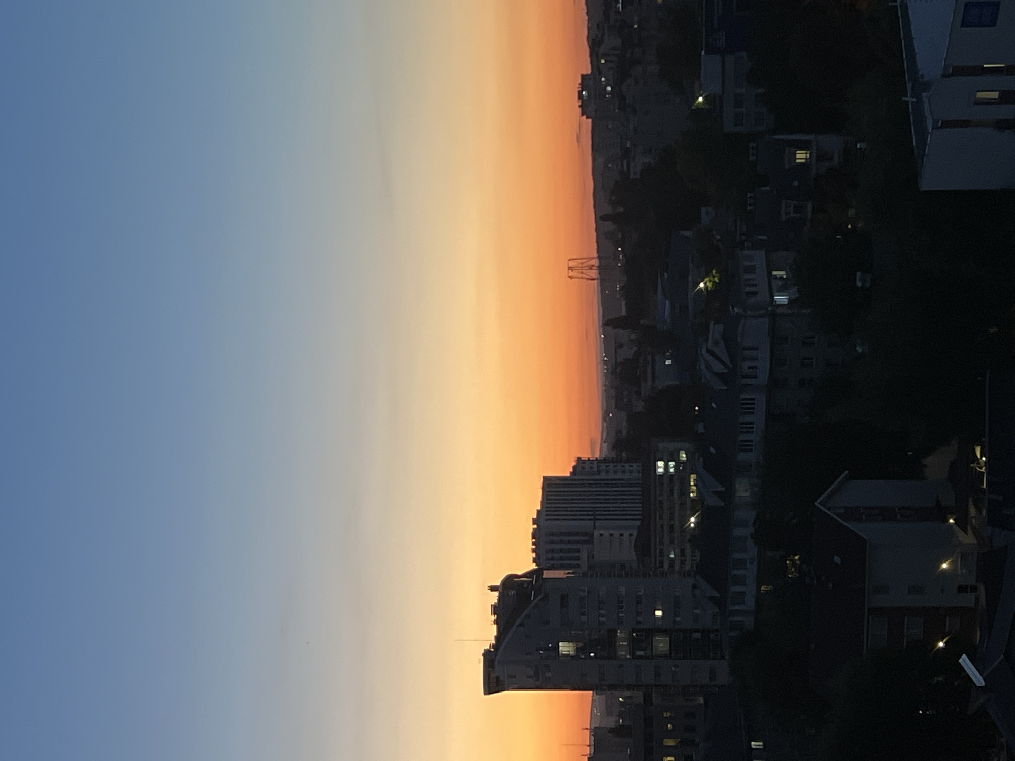 The image shows a cityscape at dusk in Chișinău, Moldova. The sky transitions from a clear blue at the top to a warm orange and yellow gradient near the horizon, indicating sunset. Several buildings are visible, including a tall, modern, curved building with illuminated windows in the foreground to the left. Behind it, other buildings of varying heights and architectural styles are present, some with lights on, suggesting evening activity. Trees and other foliage are interspersed throughout the city, appearing as dark masses against the lighter sky. In the distance, there's a glimpse of a construction or industrial structure with a distinctive lattice design. The overall scene depicts a typical urban environment during twilight.