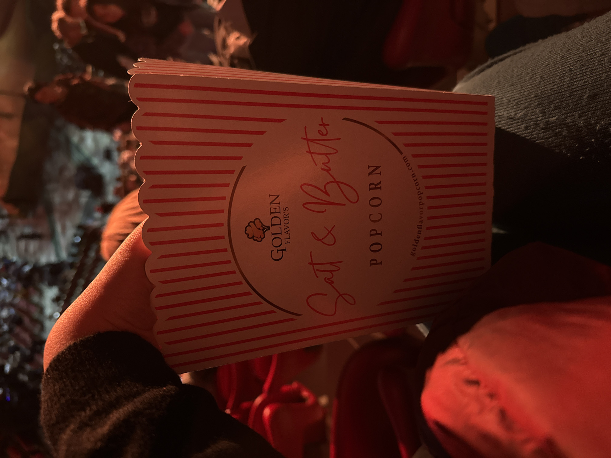 A person is holding a box of "Golden Flavor's Salt & Butter Popcorn" in what appears to be a dimly lit interior space. The box has a red and white striped design. The person's hand and part of their dark sleeve are visible. Seating and other people are in the background, slightly out of focus. The location is not identifiable as the city of Sofia, Bulgaria, based on the visual information available in the image.