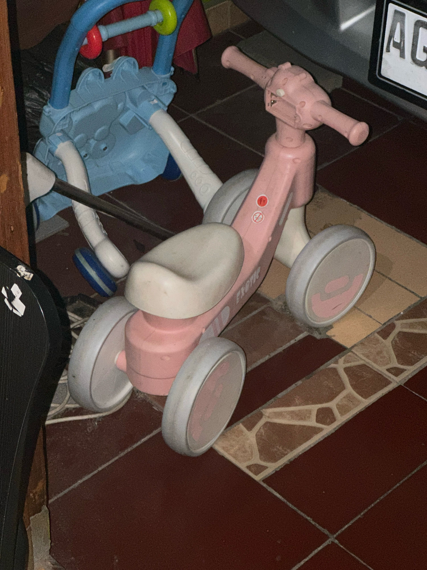 The image depicts a pink and white children's ride-on toy, possibly a balance bike, situated on a tiled floor. The toy has a pink frame, handlebars, and seat, with white wheels. A blue and white structure, possibly another toy or baby equipment, is partially visible behind it. The floor is made of reddish-brown tiles with some lighter beige tiles arranged in a mosaic pattern. In the background, a sign with the letters "AG" is visible. The setting appears to be an indoor environment, possibly a home or a storage area, in Ngasem, Indonesia.