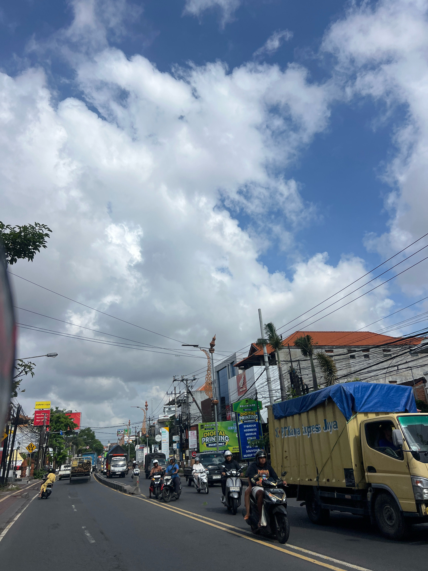The image depicts a busy street scene in Denpasar, Indonesia, under a partly cloudy sky. The foreground shows a road with vehicles including motorcycles and trucks. Several people are seen riding motorcycles. Buildings line the street, featuring signage and various architectural styles. The overall impression is one of urban activity and everyday life in a tropical climate.