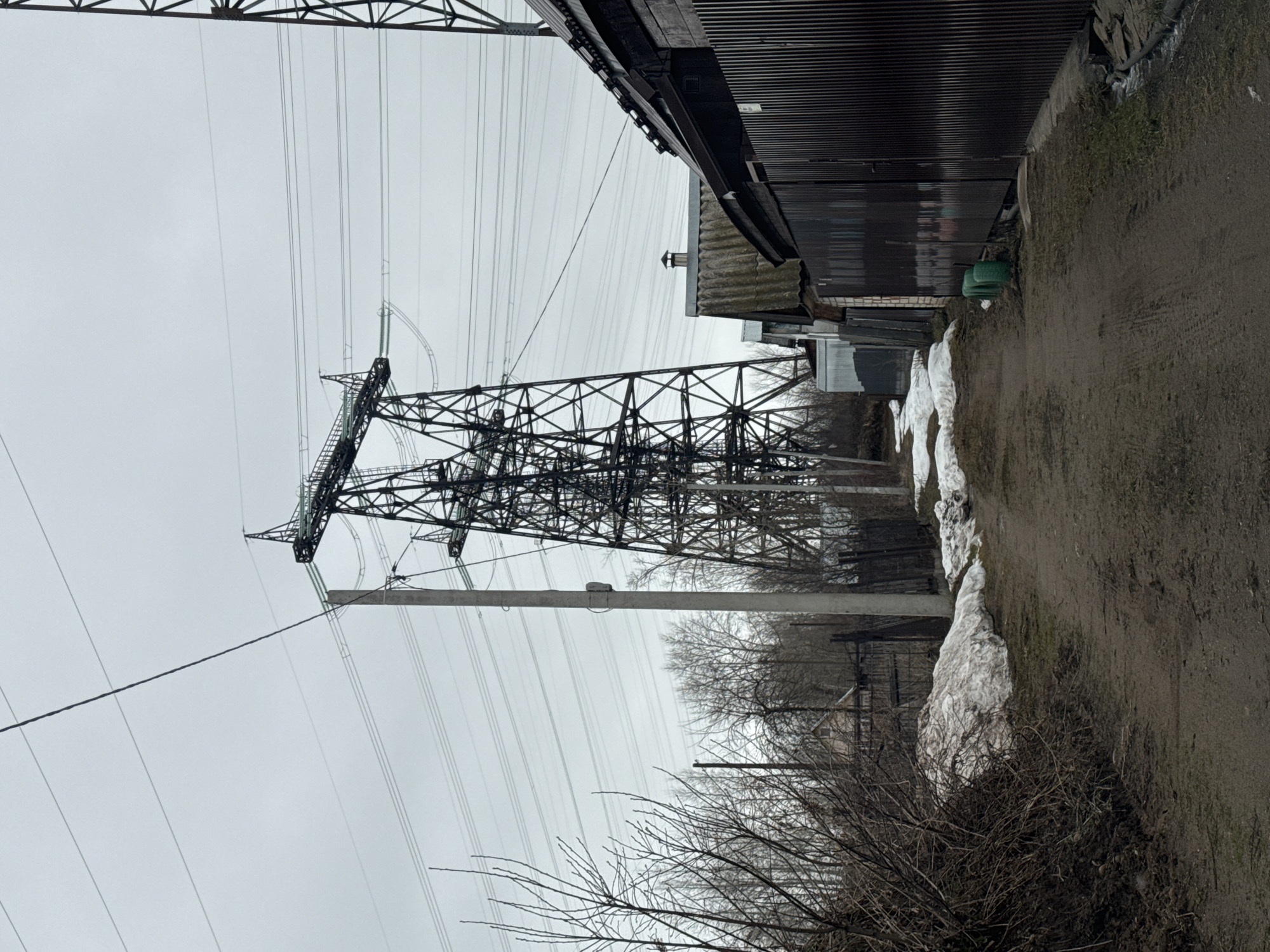 The image shows an electrical transmission tower and lines adjacent to a structure, possibly a building or shed, in Chaikovsky, Russia. The ground is dirt and shows some patches of melting snow. The sky is overcast. There are bare trees and bushes in the foreground and between the tower and building. The transmission tower is a complex lattice structure, and the power lines stretch out across the sky. The building is dark, with corrugated metal siding.